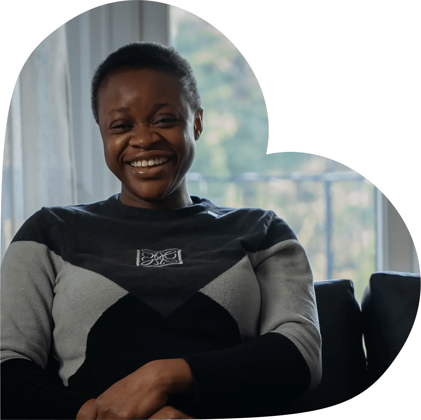 Smiling woman with short hair wearing a black, grey, and white sweater, sitting indoors near a window.