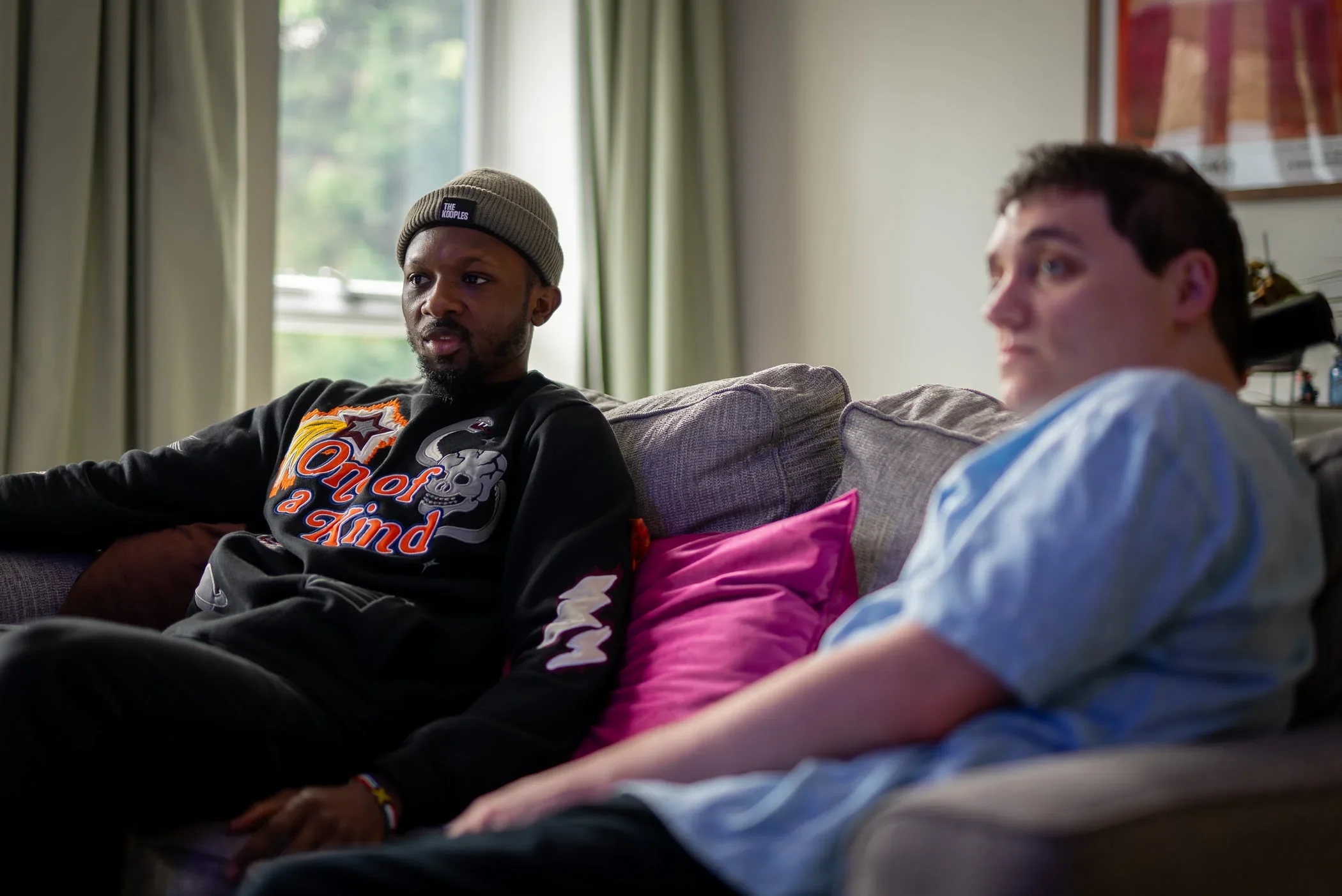 Two men sitting on a living room couch, one with a serious expression wearing a beanie and a black sweatshirt with colourful text and graphics, and the other wearing a light blue t-shirt, looking ahead, with a pink pillow behind the first man.