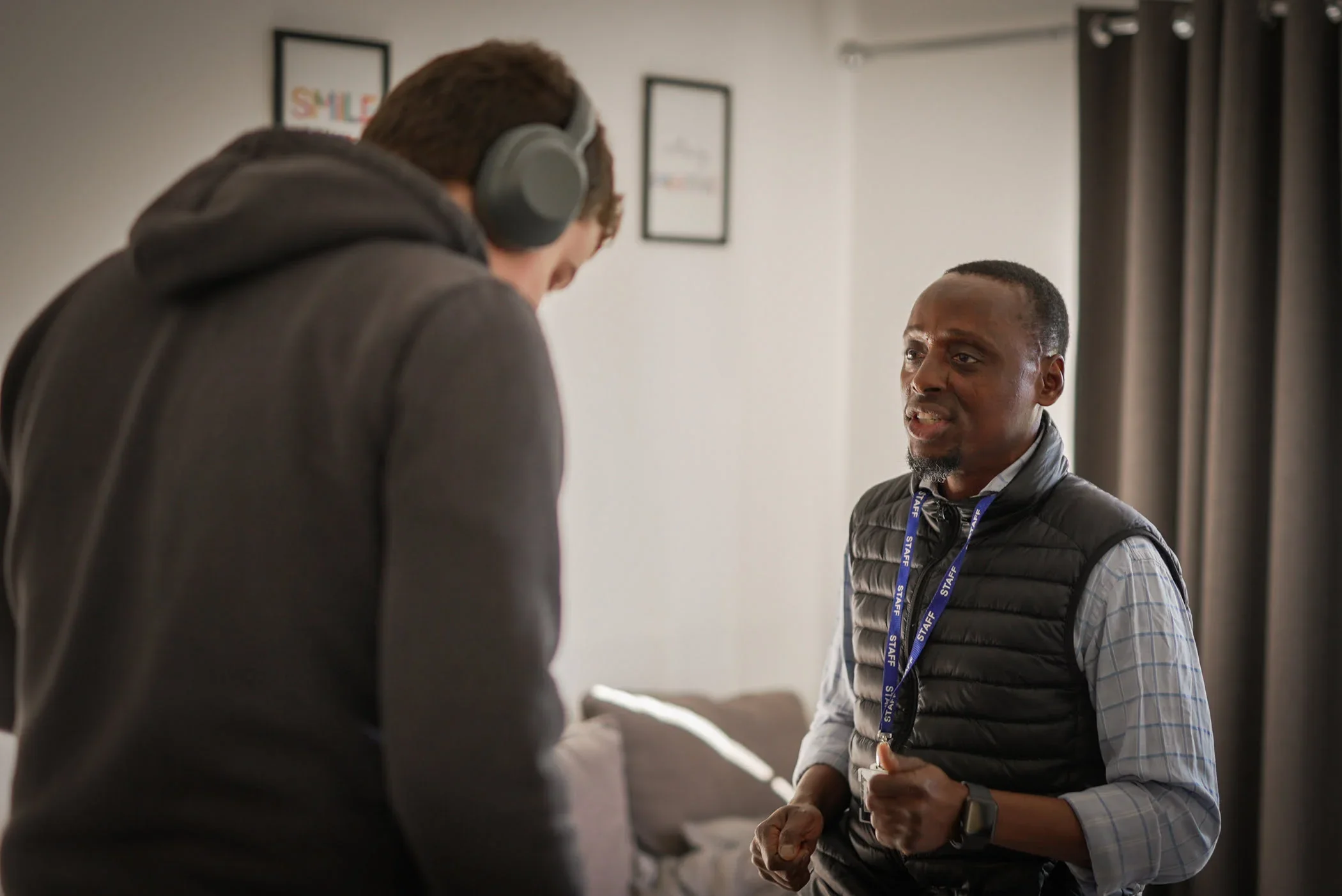 Two men engaged in conversation indoors; one man with headphones and casual attire, the other with a lanyard, vest, and a checkered shirt, expressing emotions.