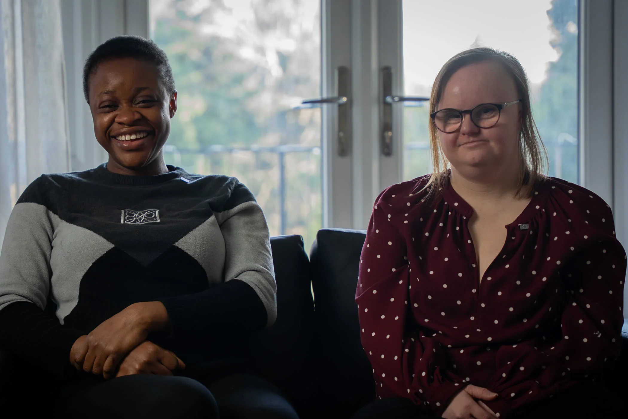 Two women sitting on a black couch in front of large glass doors, smiling and looking at the camera. The woman on the left has dark skin, short black hair, and is wearing a patterned gray and black sweater. The woman on the right has light skin, long light brown hair, glasses, and is wearing a maroon blouse with white polka dots.