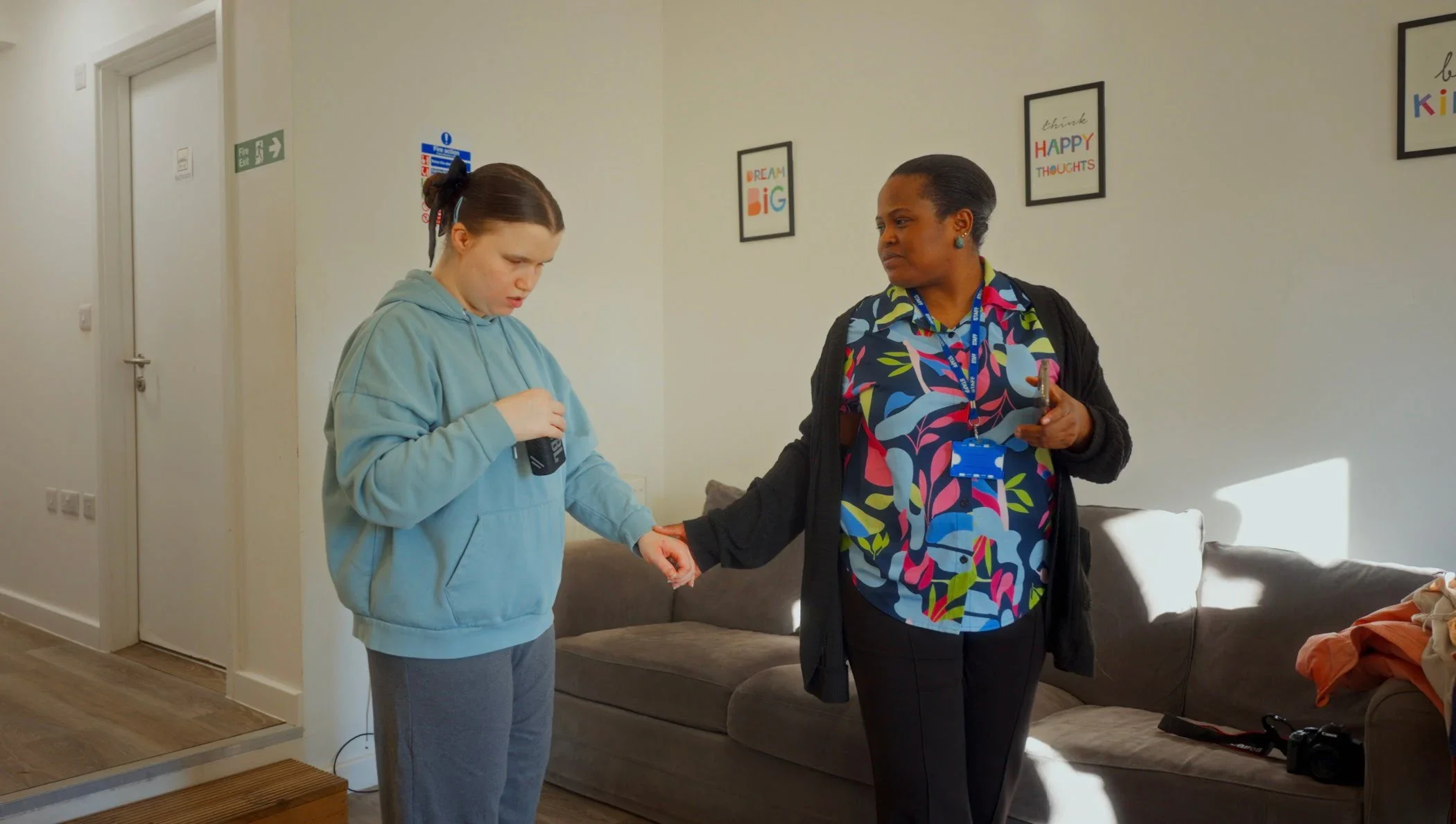 Two women standing in a living room, one young with a hoodie, holding a device, and an older woman with attached lanyard, holding a phone and touching the young woman's arm. Wall decorations include framed quotes.