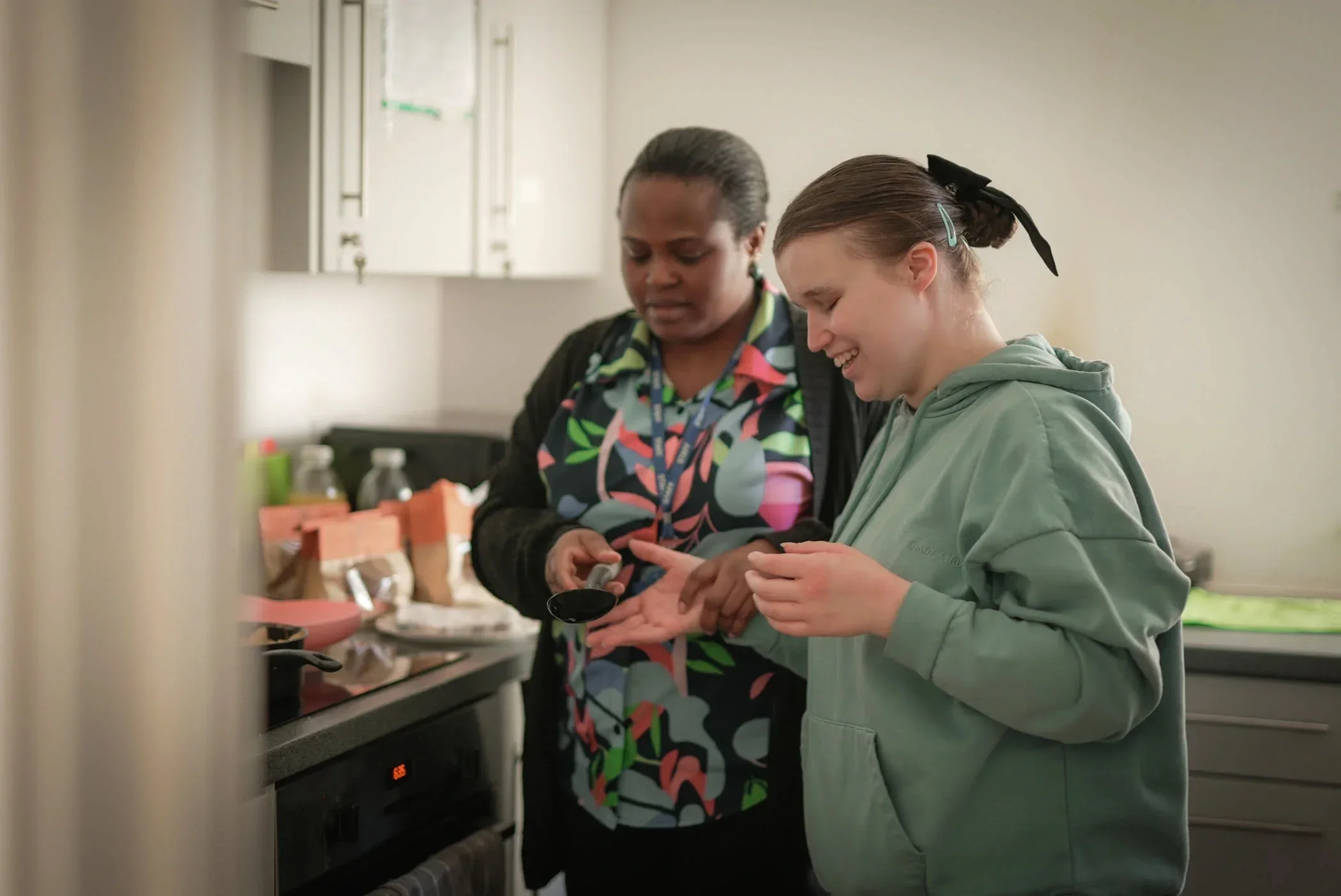 A young girl with a black ribbon in her hair and an older woman at a kitchen stove, smiling and using a spice jar.