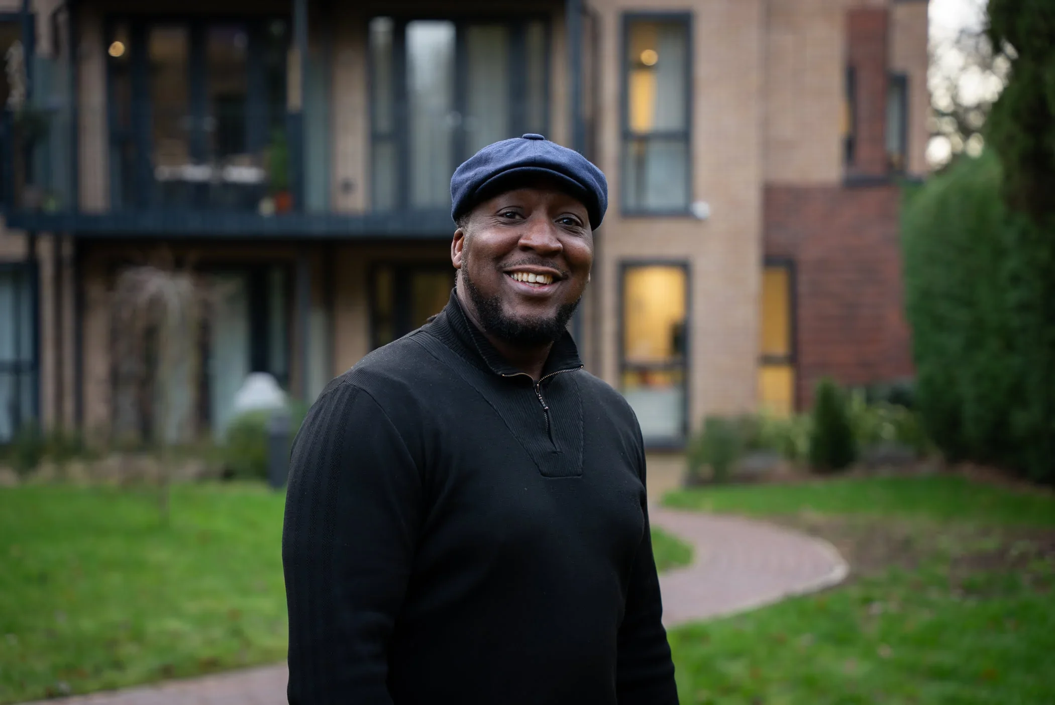 A smiling man wearing a black sweater and a navy flat cap standing outdoors in front of apartment buildings and a grassy area.