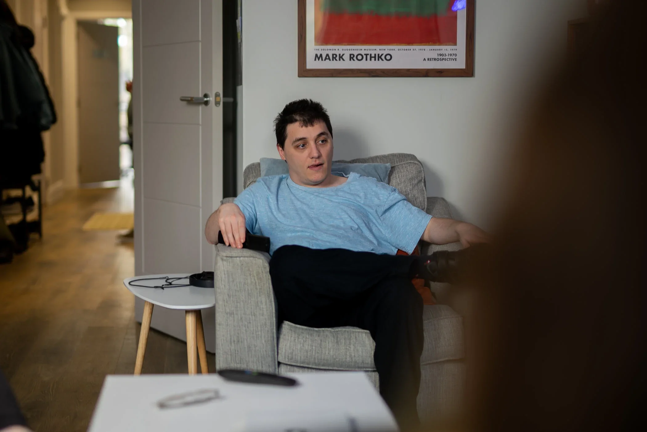 Young man sitting on a beige armchair in a living room, wearing a blue shirt, with a serious expression, holding a TV remote. There is a small table with a TV remote and other items beside him, with a poster of artist Mark Rothko hanging on the wall behind him.