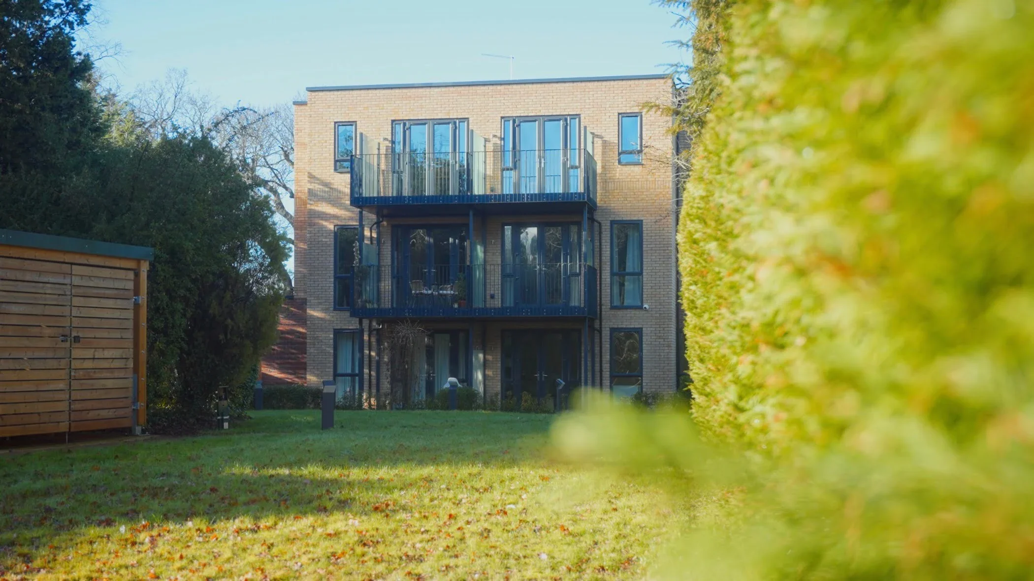 A three-story brick apartment building with large glass balconies, surrounded by a green lawn, tall hedges on the right, and trees on the left, under a clear blue sky.