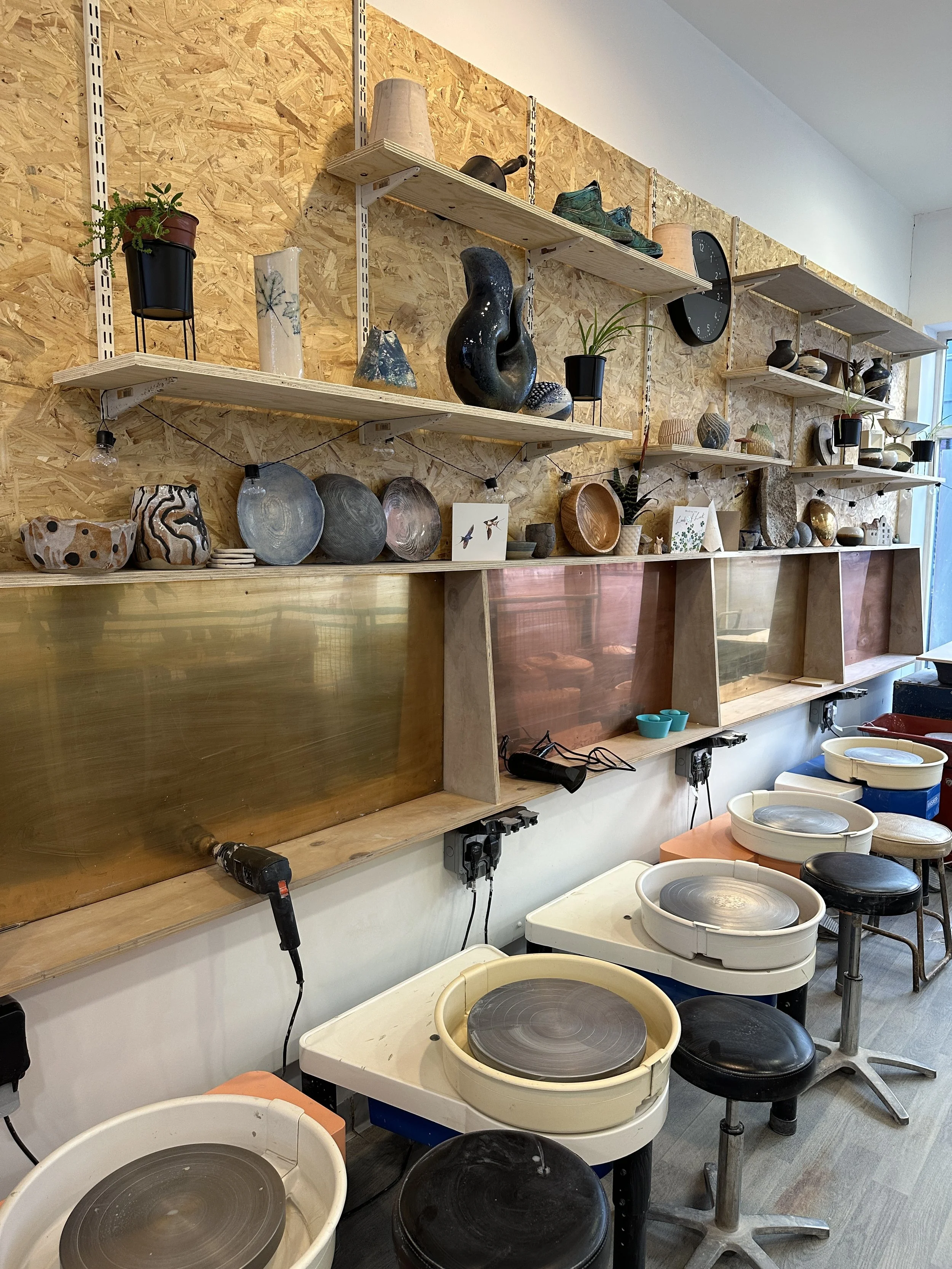 Four pottery wheels lined up in a workshop with a wooden wall and a window behind them.