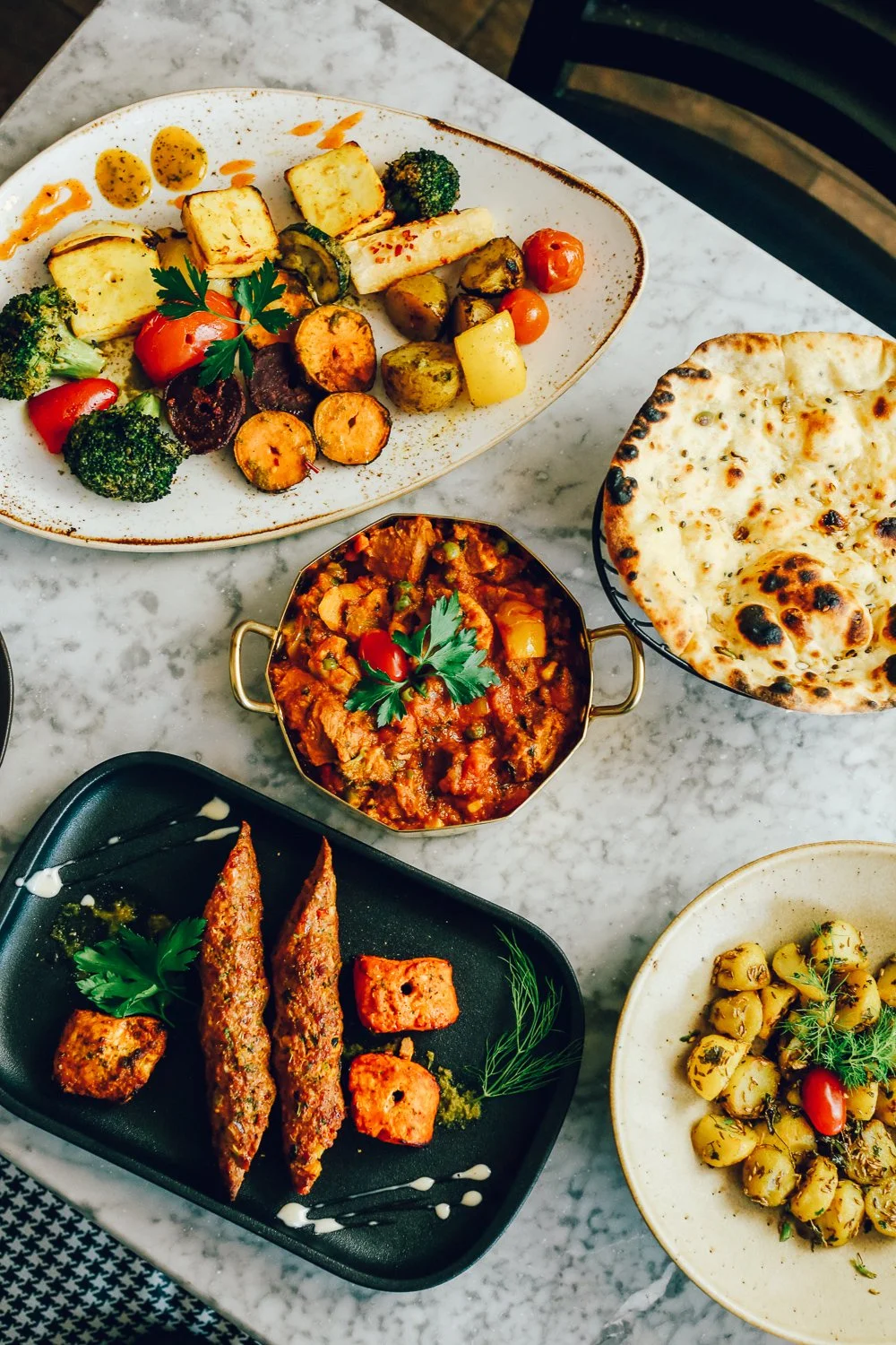 An assortment of Indian dishes on a marble table, including naan bread, a vegetable curry, grilled kebabs, roasted vegetables, and chickpea salad.