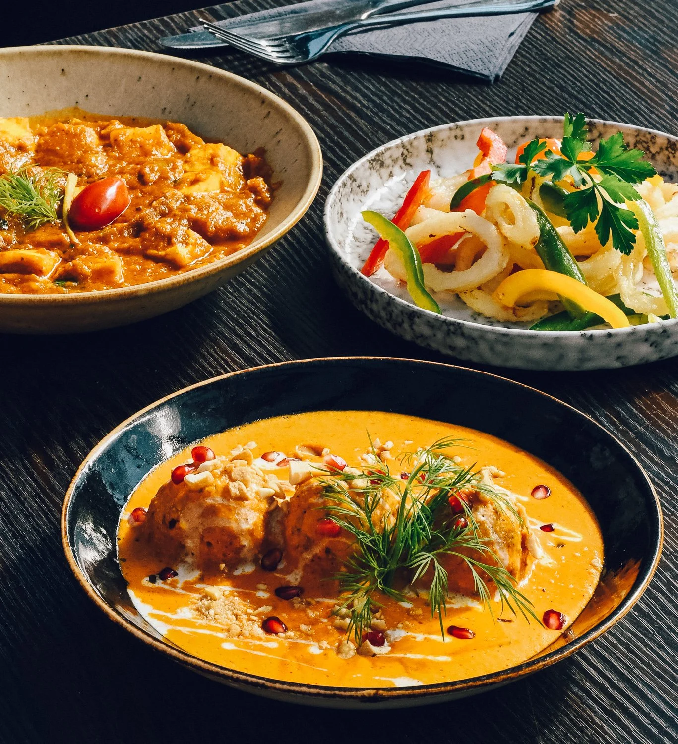Three bowls of Indian cuisine on a dark wooden table: a bowl of butter chicken with garnish, a bowl of stir-fried vegetables, and a bowl of chicken in an orange curry sauce with pomegranate seeds and herbs.