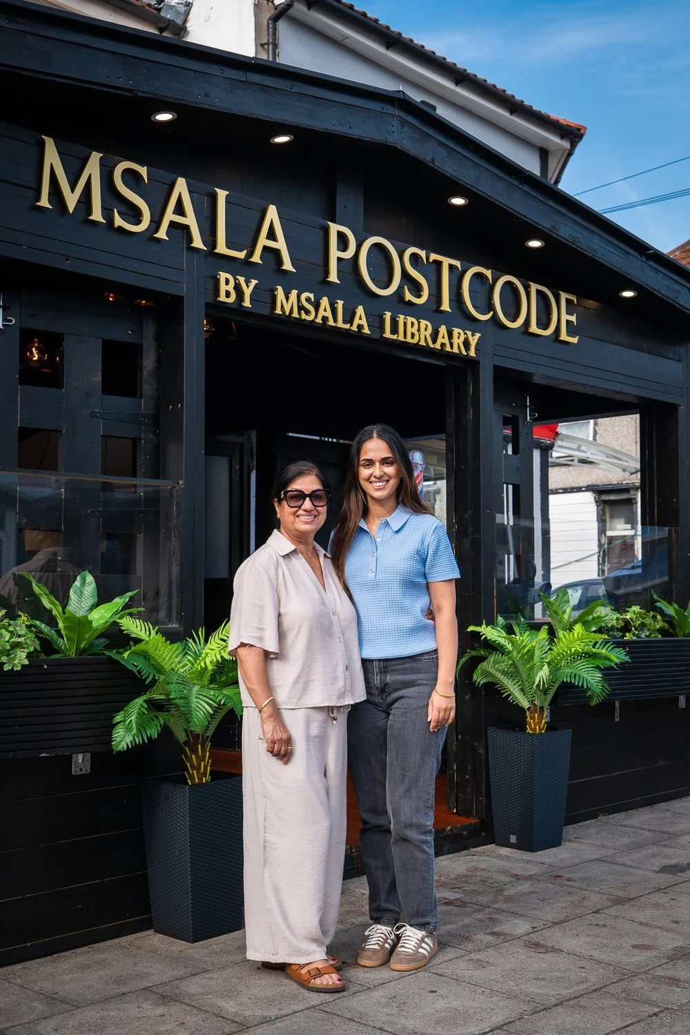 Two women standing in front of a building with a sign that reads 'MSALA POSTCODE BY MSALA LIBRARY,' smiling at the camera, surrounded by potted plants.