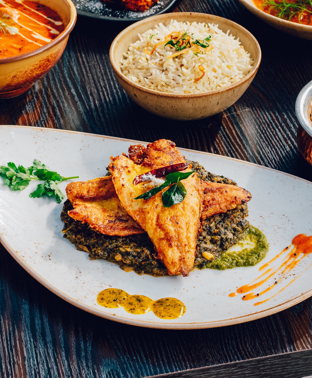 Plate of fried fish atop cooked spinach, garnished with herbs and sauces, with bowls of rice and lentil stew in the background.