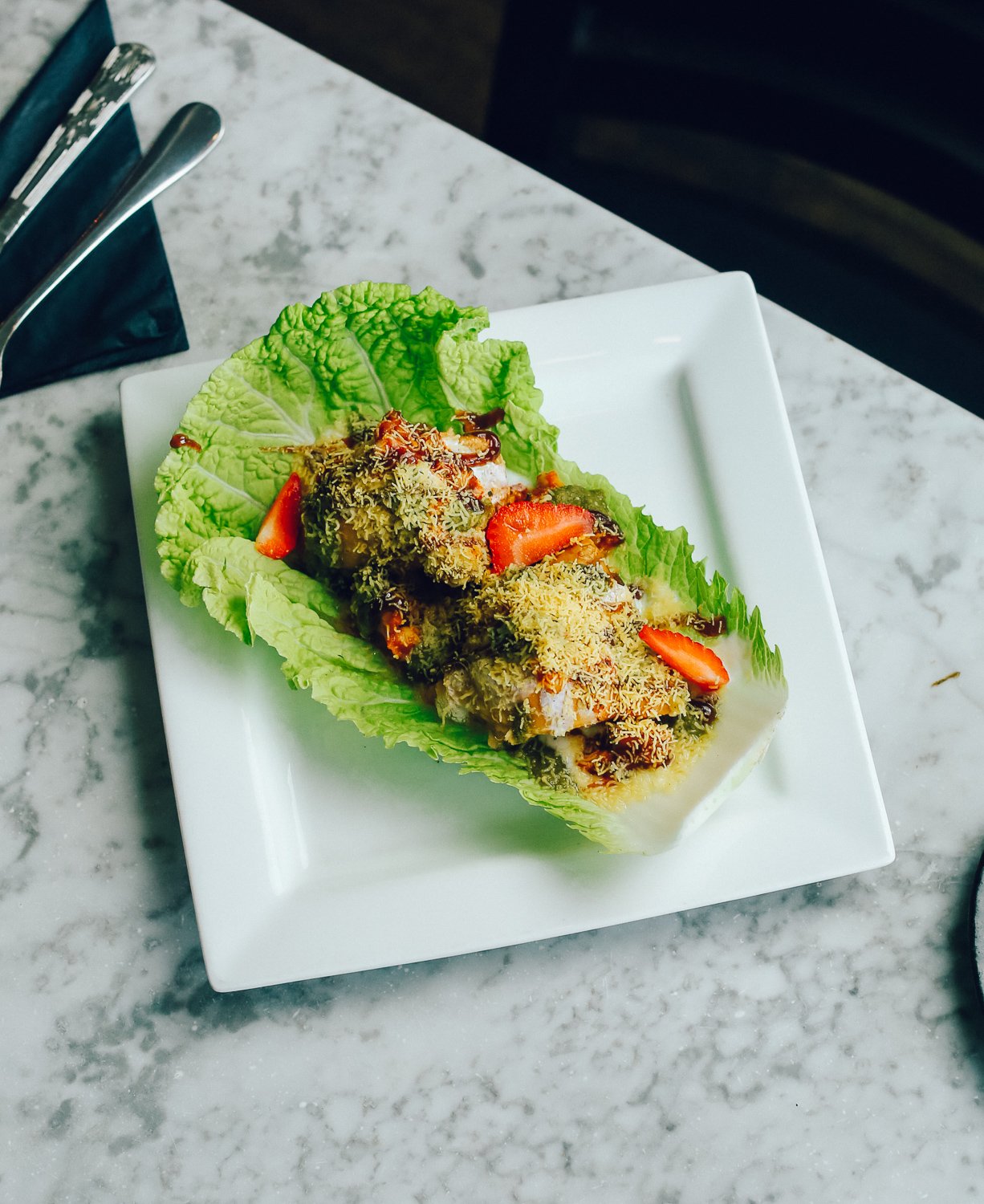Lettuce leaf topped with sliced strawberries, shredded cheese, and drizzled with sauce on a white square plate.