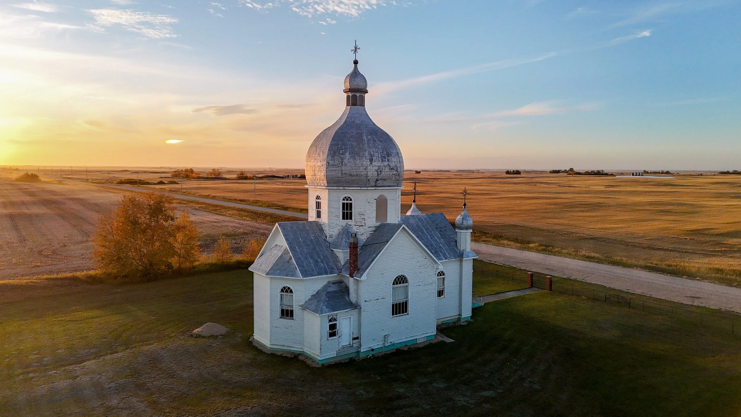 A white wooden church with a large onion dome and smaller domes, surrounded by farmland at sunset.