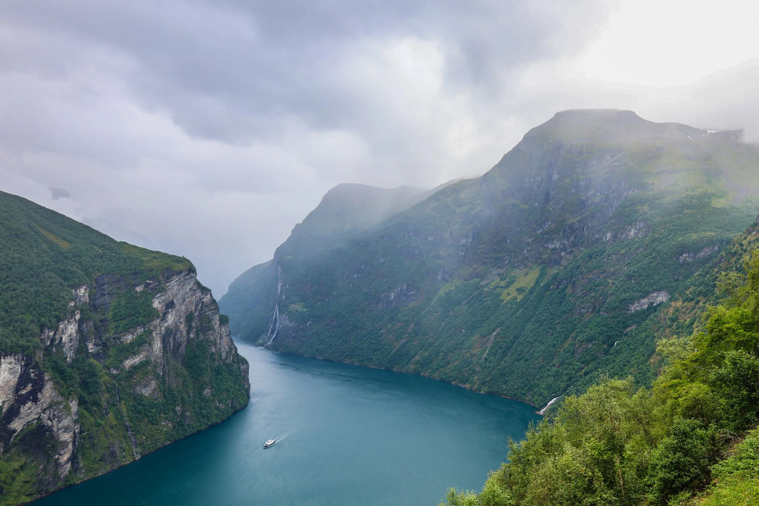 A scenic view of a narrow fjord with steep, lush green mountains on either side, a boat sailing in the water, and misty clouds overhead.