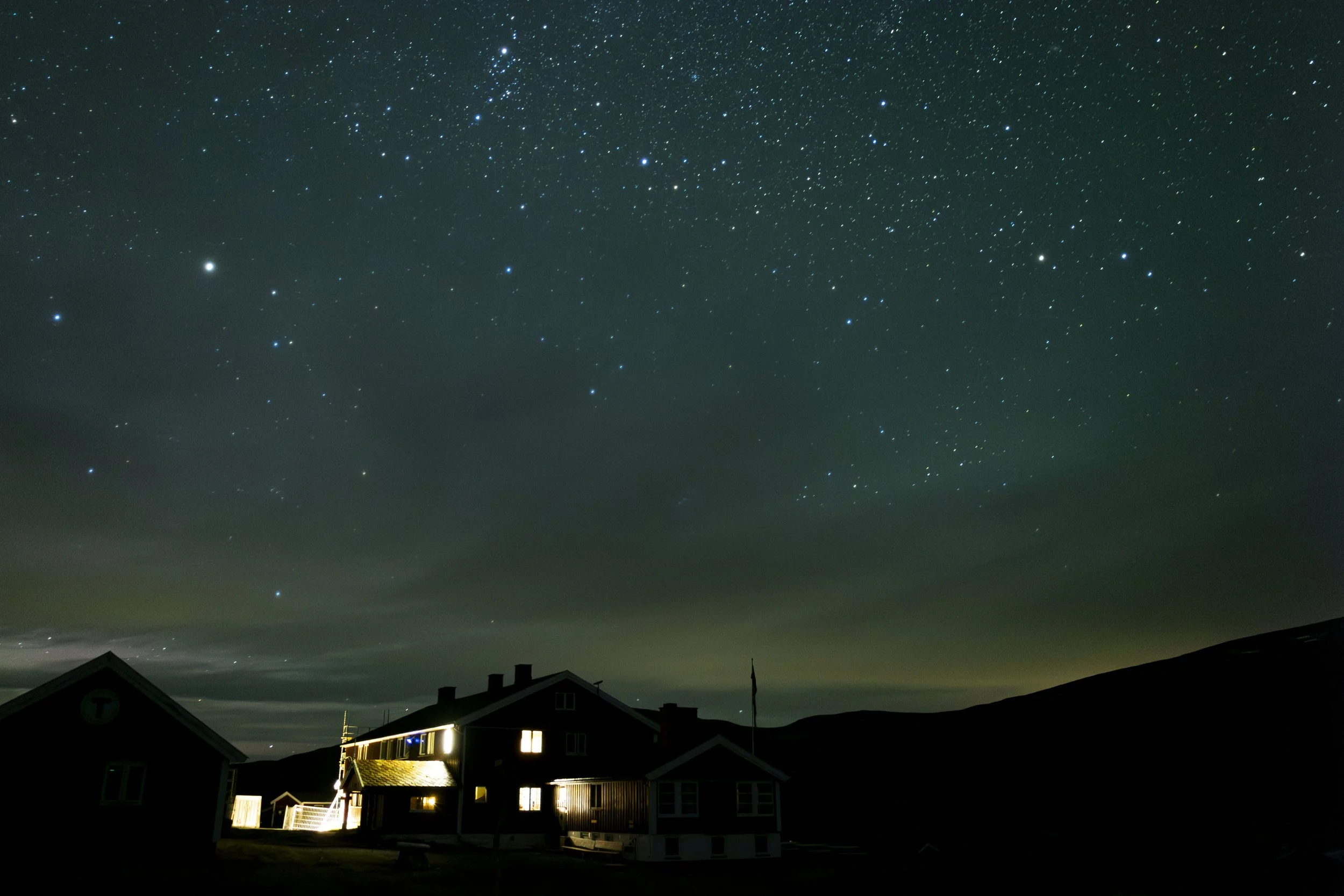 Night sky filled with stars over a dark rural landscape with houses and hills.