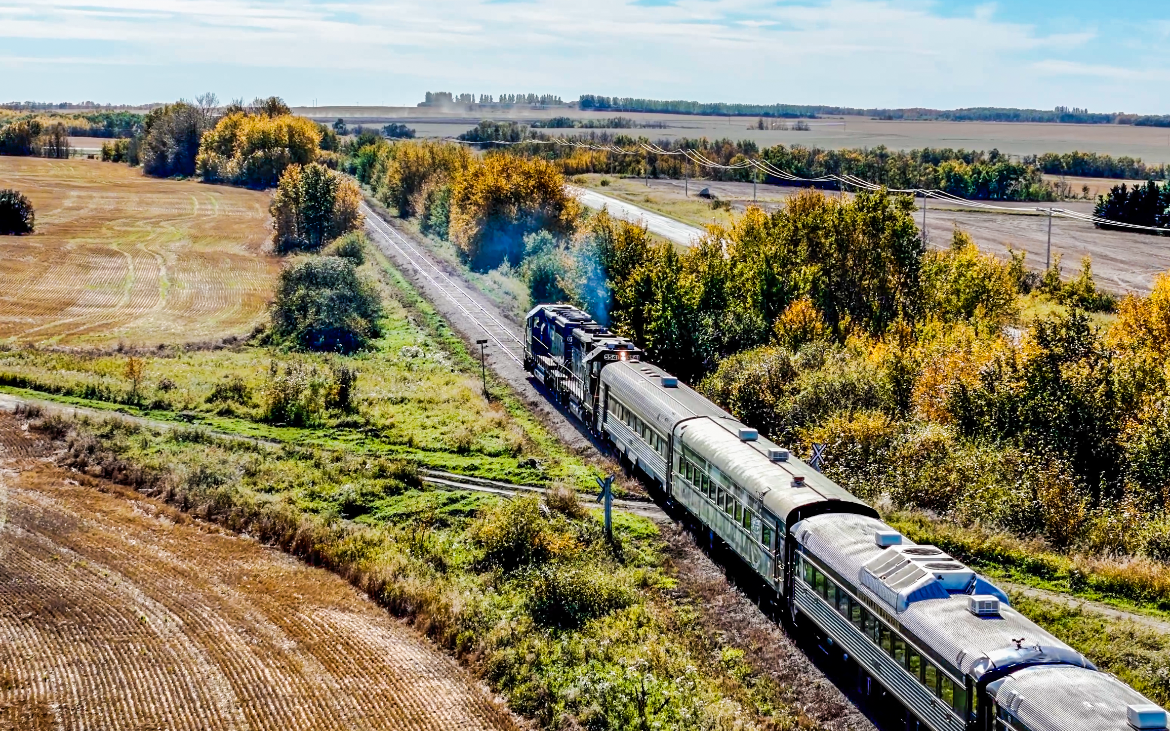 A train moving through a rural landscape with fields and trees, under a partly cloudy sky.