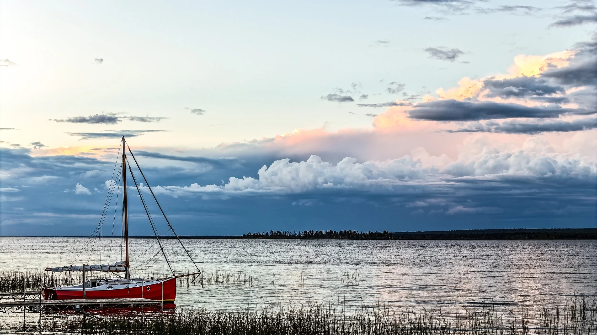 A red sailboat docked at the shore of a calm lake, with a cloudy sky and distant forest in the background