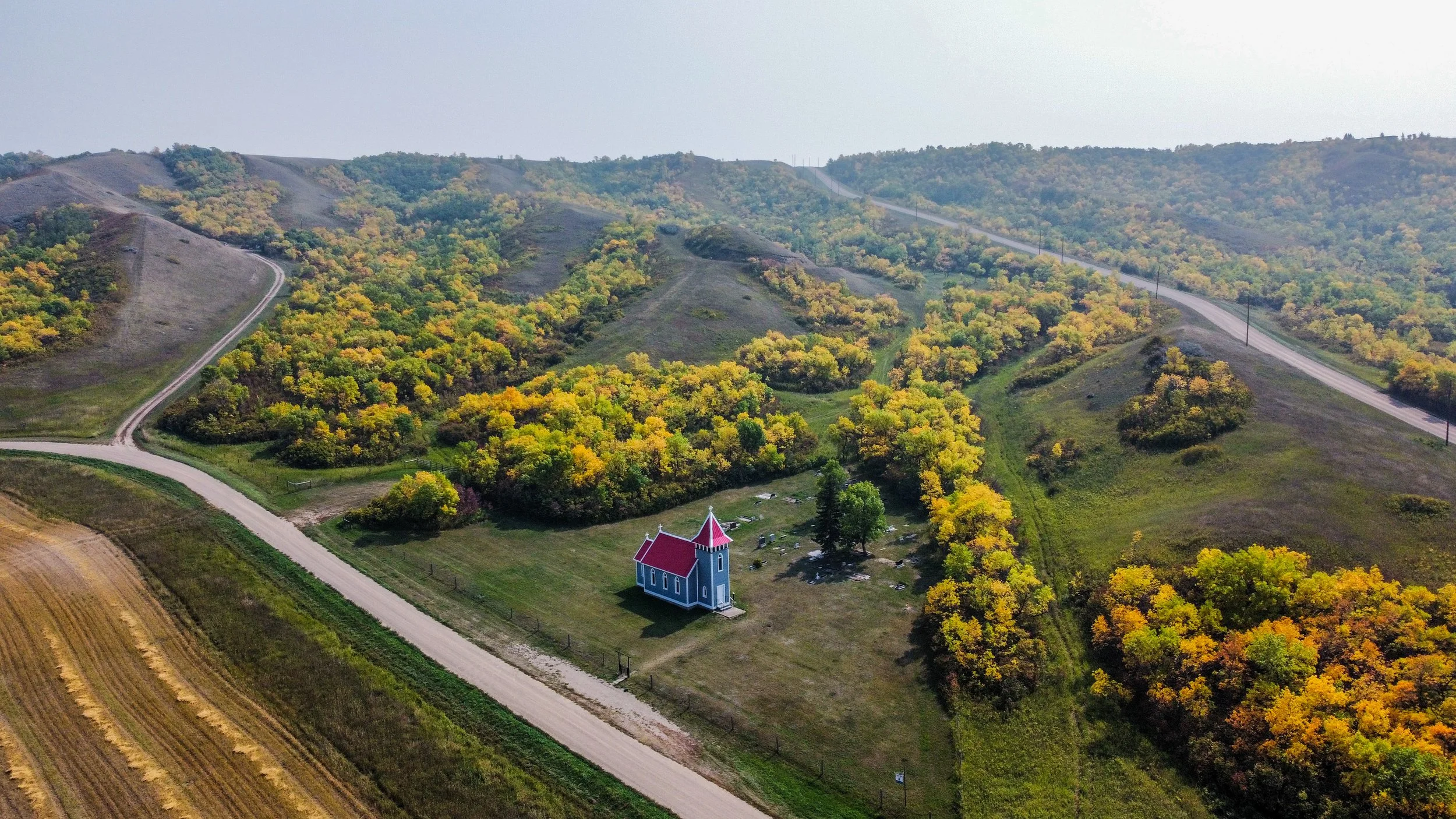 An aerial view of a small blue church with a red roof and steeple, surrounded by green trees with hints of orange and yellow, situated on a grassy area near a dirt road in a hilly landscape.