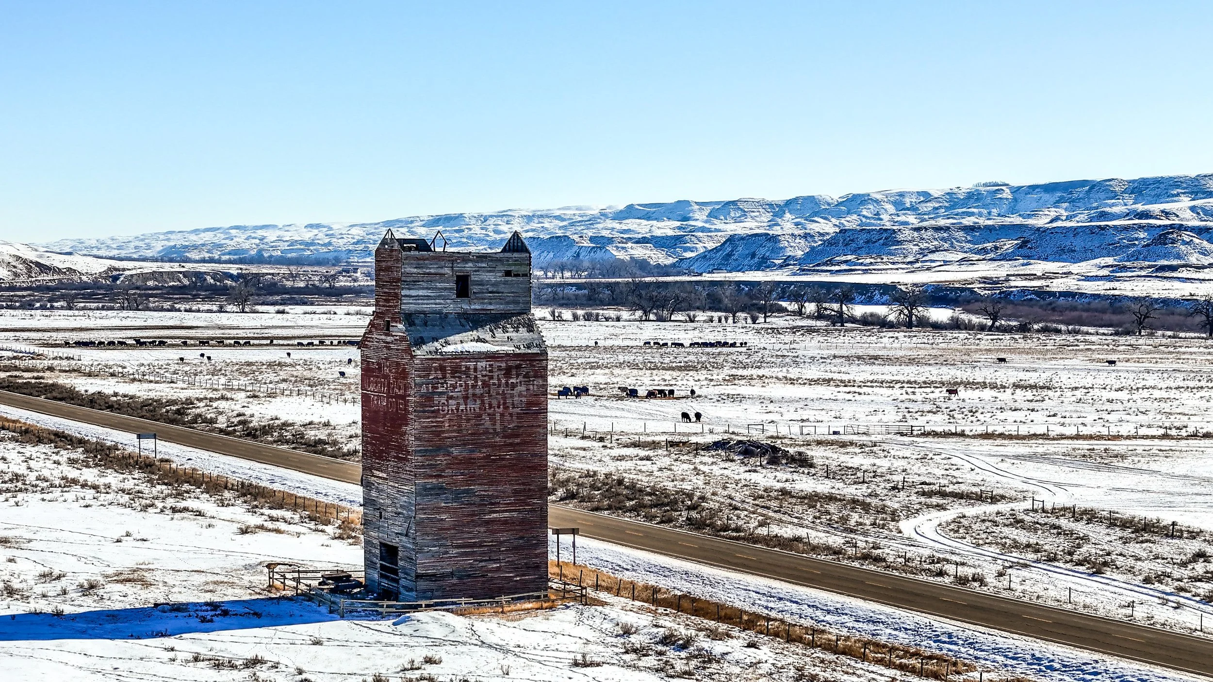 A weathered, old wooden grain elevator building in a snowy rural landscape with mountains in the background, a road in the foreground, and cattle grazing in the distance.