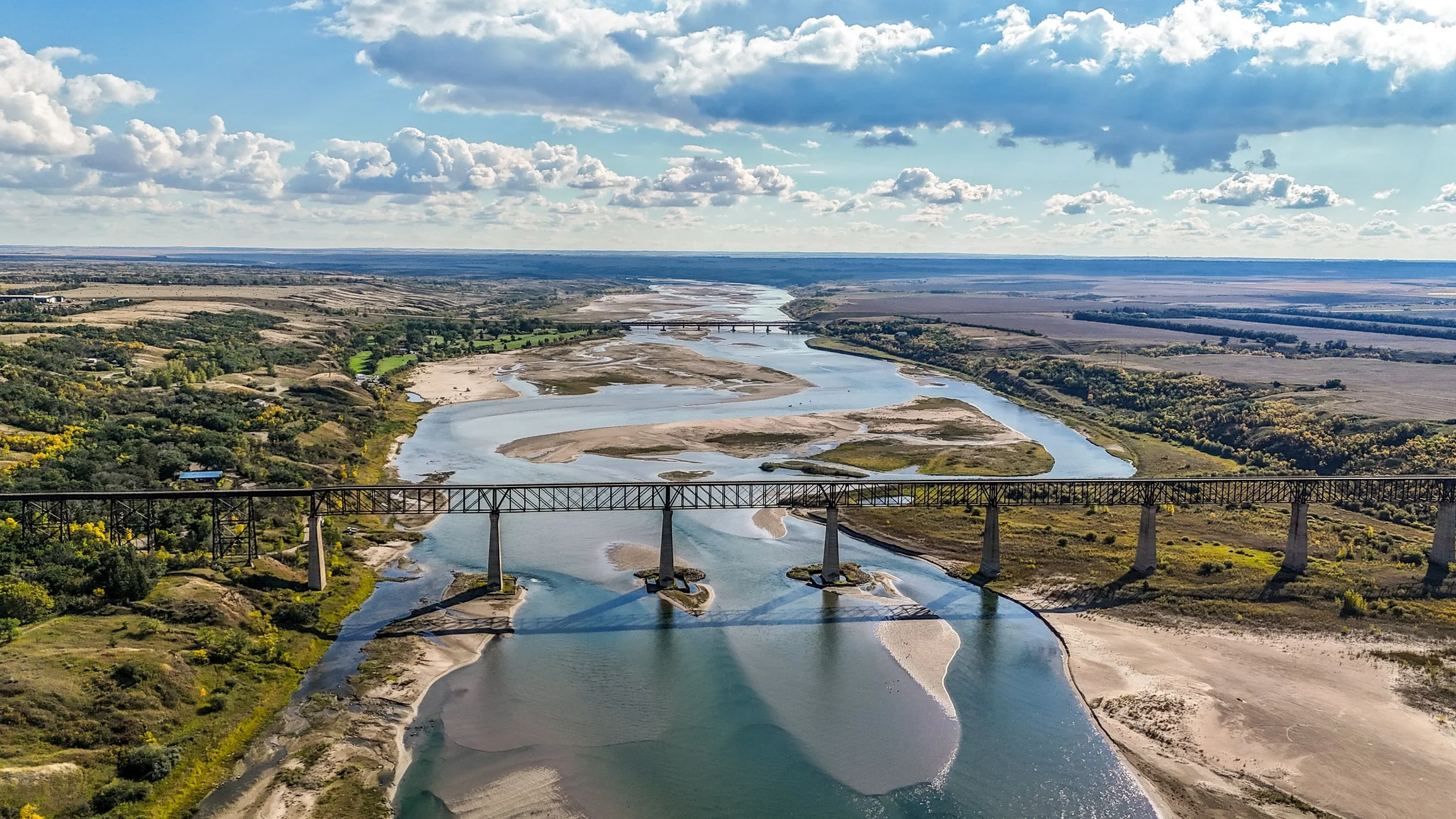 A wide river with multiple channels flowing through a landscape with patches of green and sandy areas, a bridge crossing over the river, and a sky with scattered clouds.