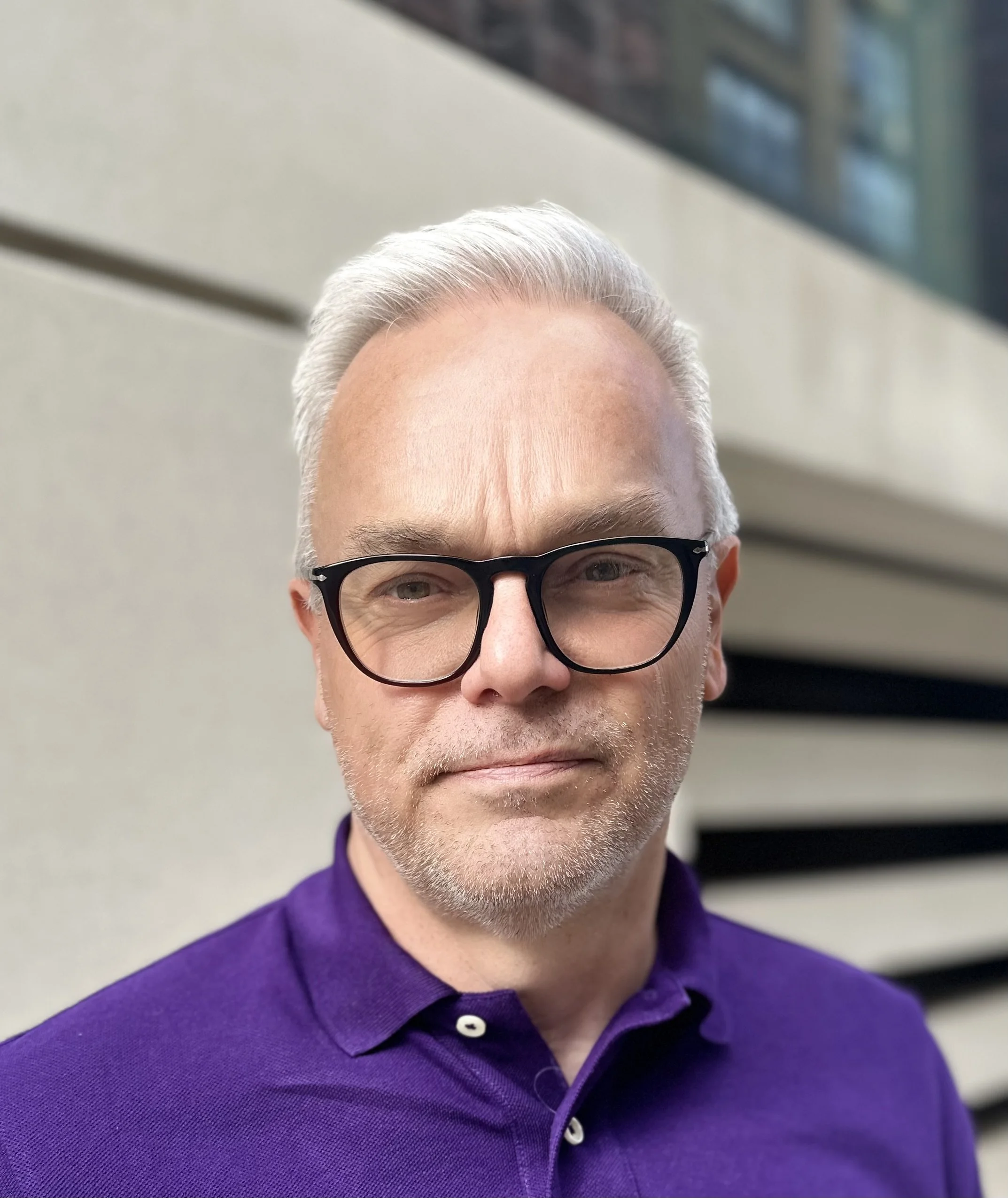 A man with gray hair and glasses wearing a purple shirt takes a selfie outside near a modern building.
