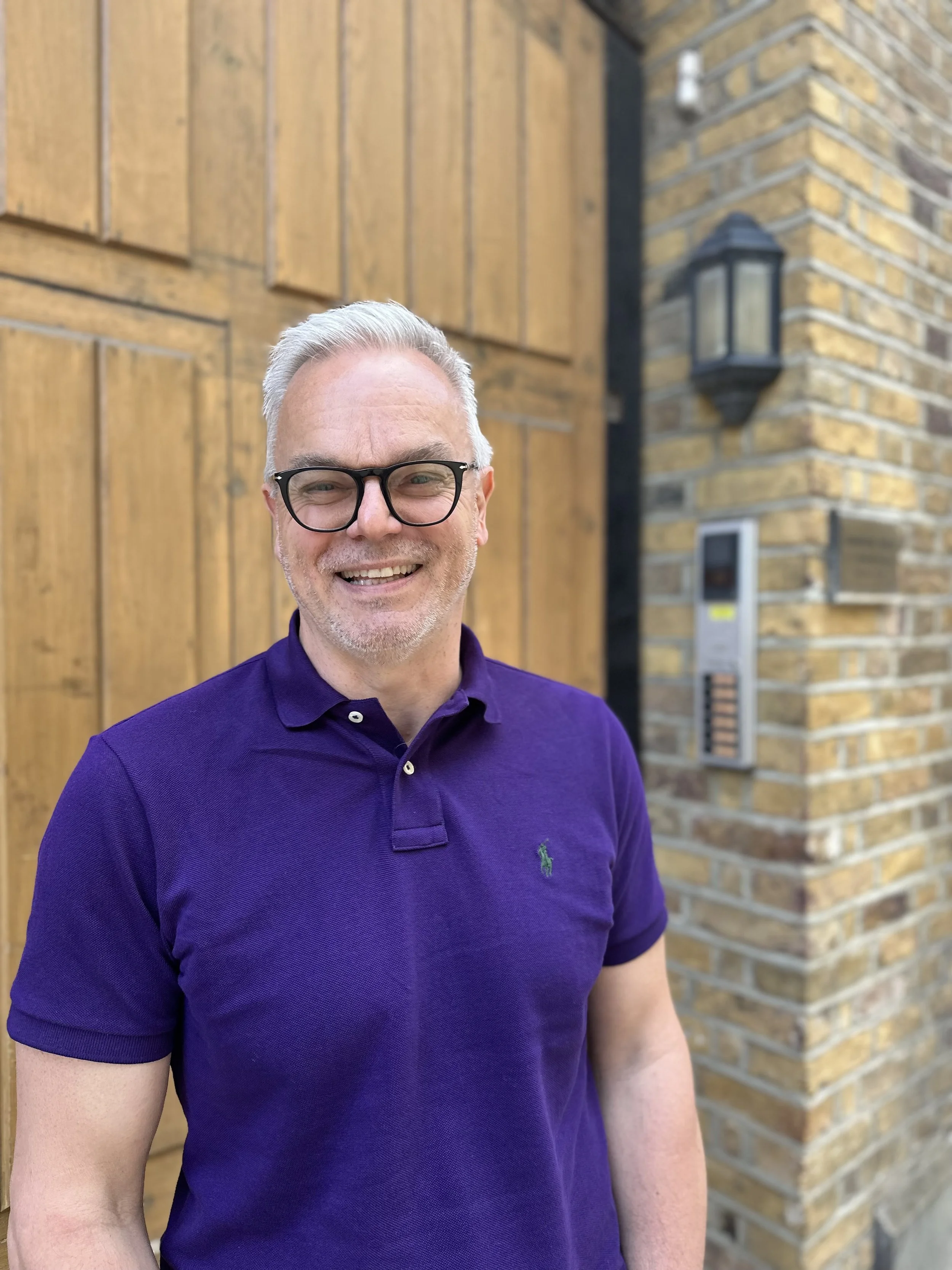 A middle-aged man with gray hair, glasses, and a beard smiling outdoors. He is wearing a purple polo shirt with a small green logo, standing in front of a wooden door and brick wall with outdoor lighting and an intercom system visible.