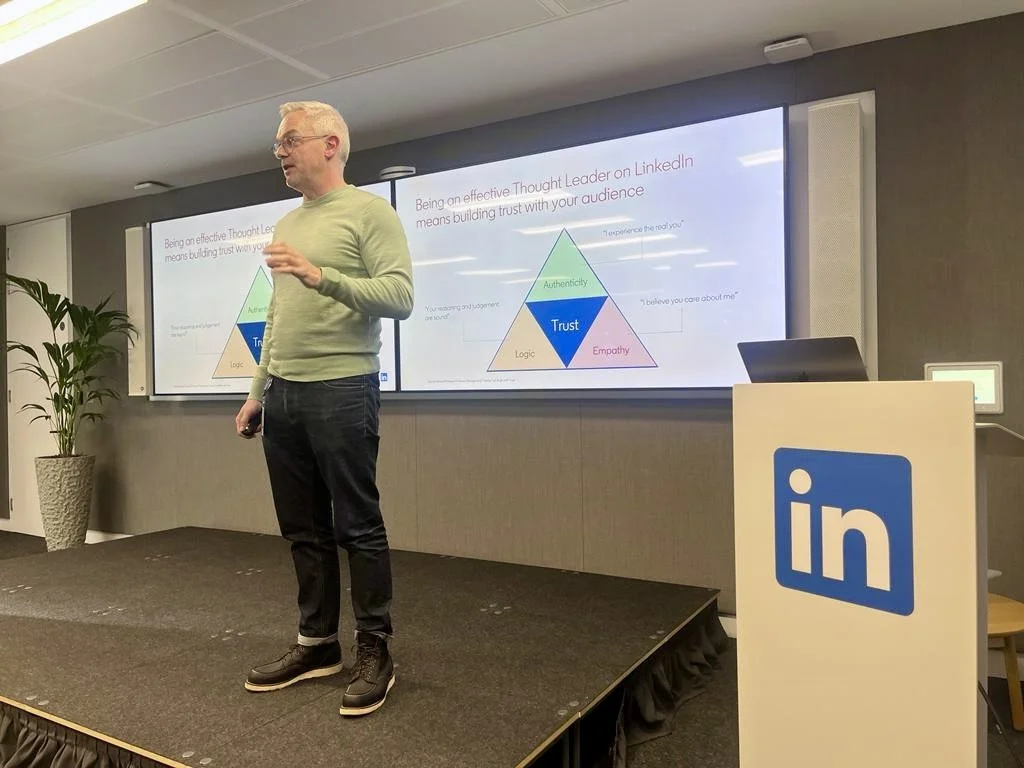 A man giving a presentation on a stage at a LinkedIn event, with a slide behind him displaying a triangle diagram about building trust through authenticity, empathy, and logic.