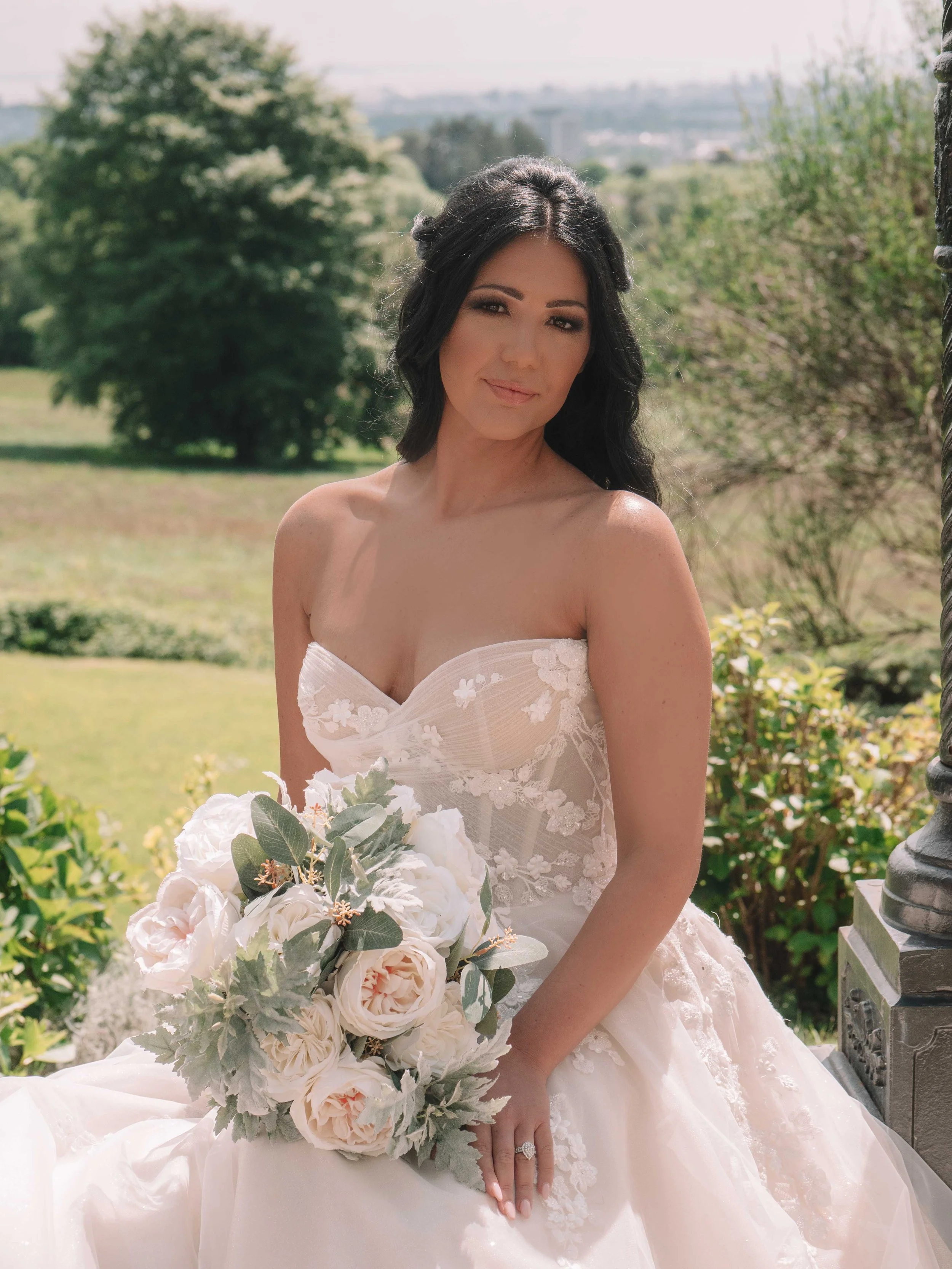 A woman in a white wedding dress holding a large bouquet of white and blush flowers, sitting outdoors on a sunny day with greenery in the background.