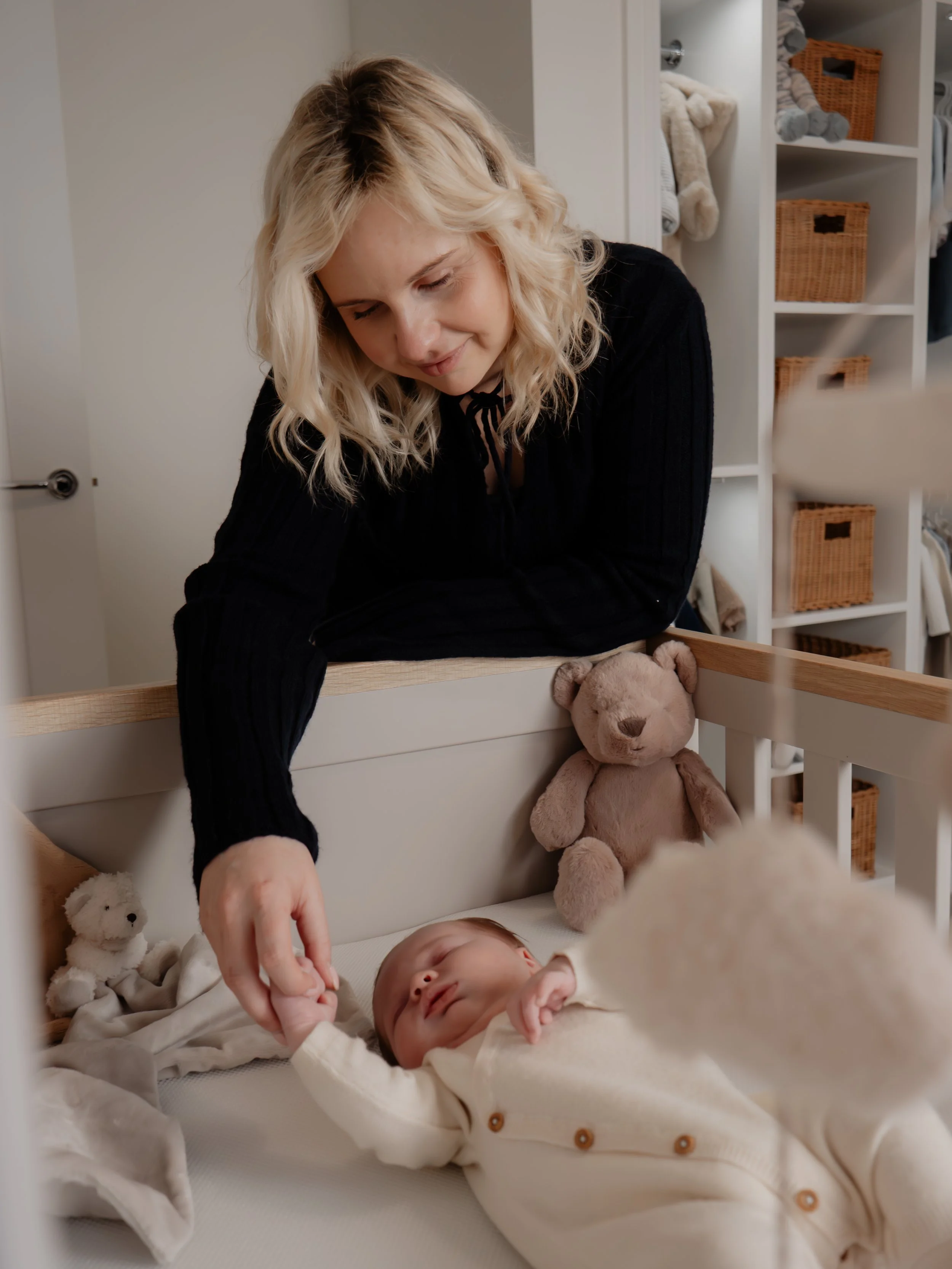 A woman watches over a sleeping baby in a crib, holding the baby's hand. The crib has teddy bears inside, with a closet in the background.