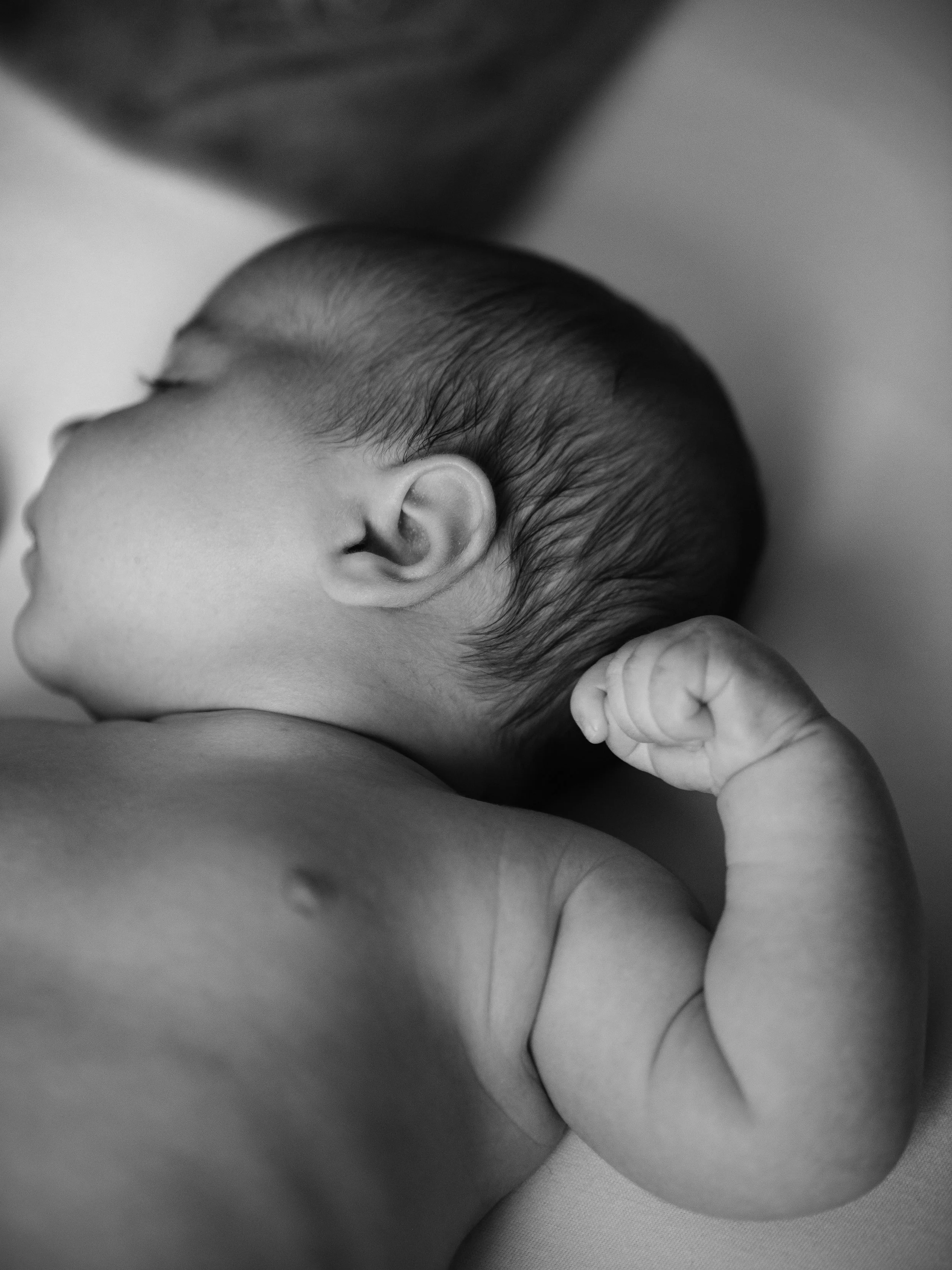 Close-up of a sleeping newborn baby with a clenched fist, lying on their side in black and white.