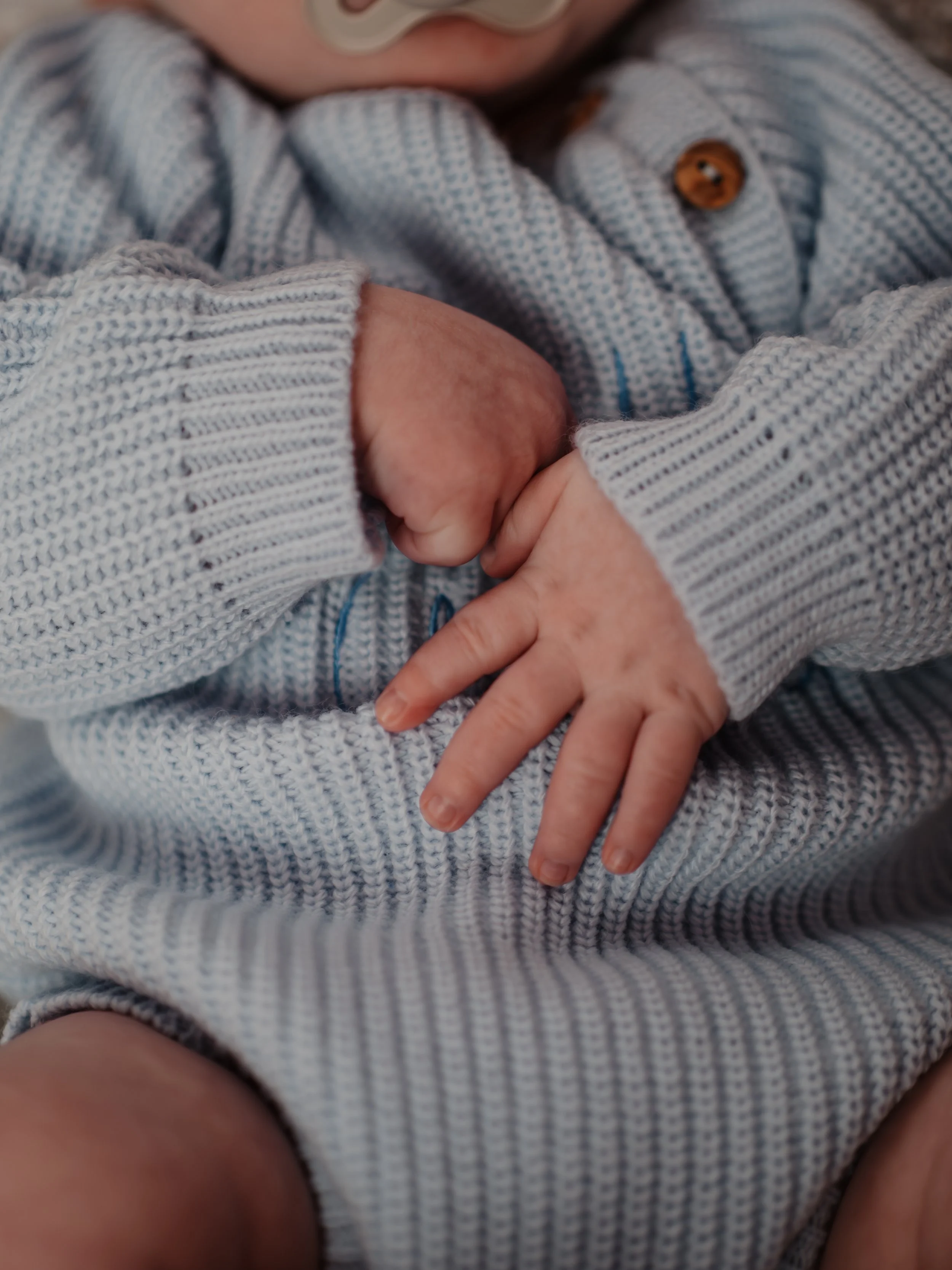 Close-up of a person's hand gently holding a child's hand, both wearing knitted sweaters, with the child's hand resting on their lap.