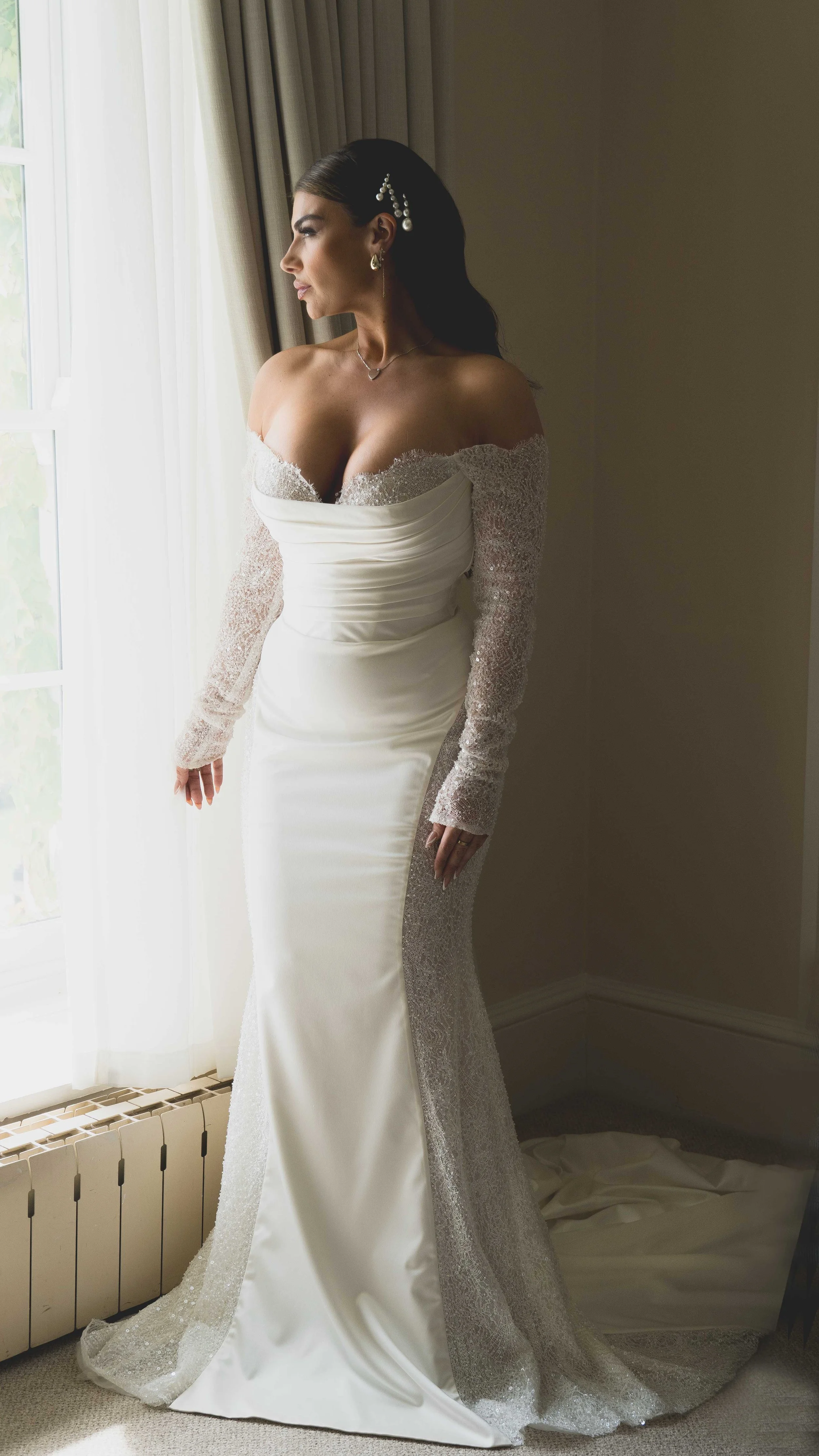 A woman in a white wedding dress with off-the-shoulder sleeves and lace details, standing by a window and looking outside.