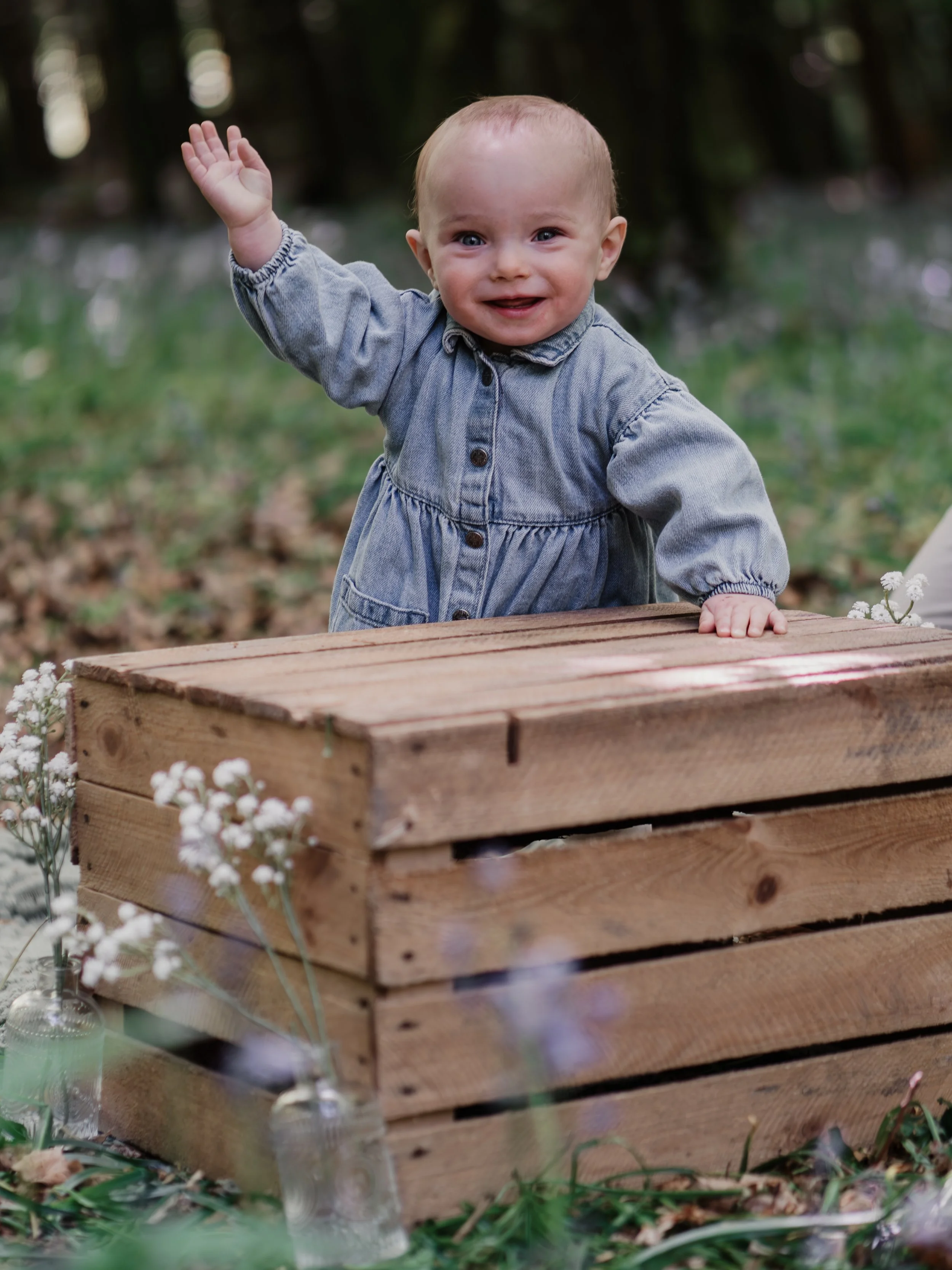 A smiling baby with blue eyes and light hair, wearing a denim dress, is leaning on a wooden crate outdoors, waving. The background is a blurred natural setting with trees and grass, decorated with small white flowers in vases.