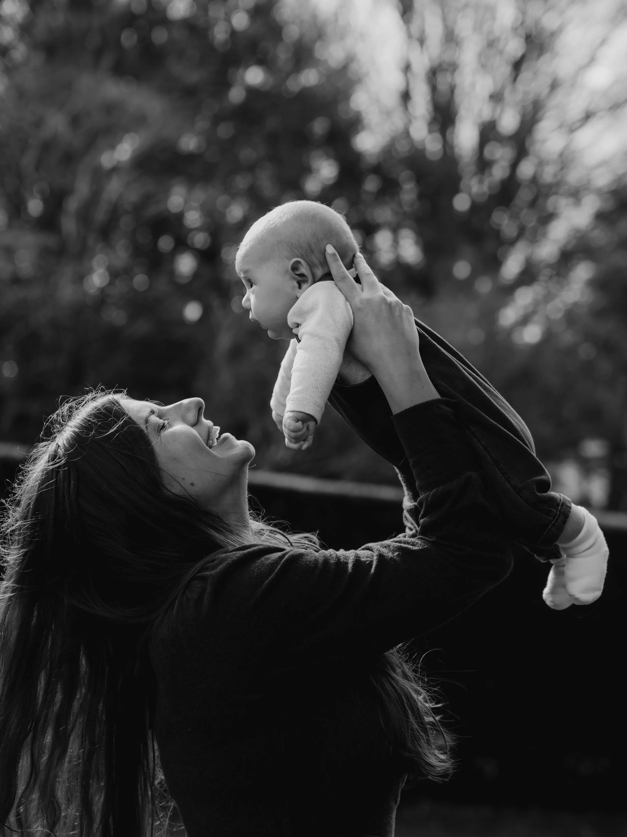 A woman smiling and holding a baby up in the air outdoors, with blurred trees in the background.