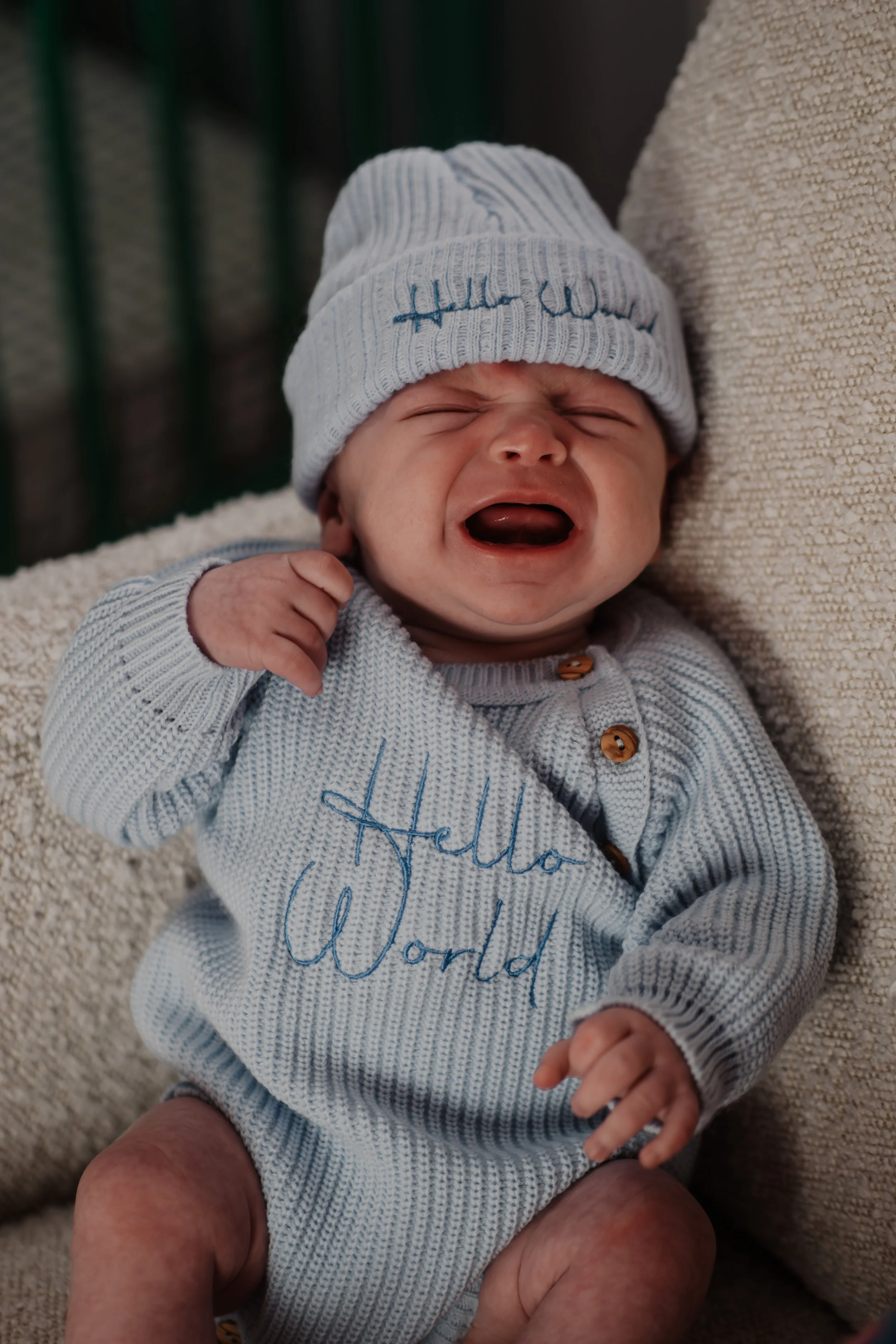 A crying baby wearing a light gray knit sweater and a matching hat with 'Hello World' embroidered on it, lying on a textured blanket.