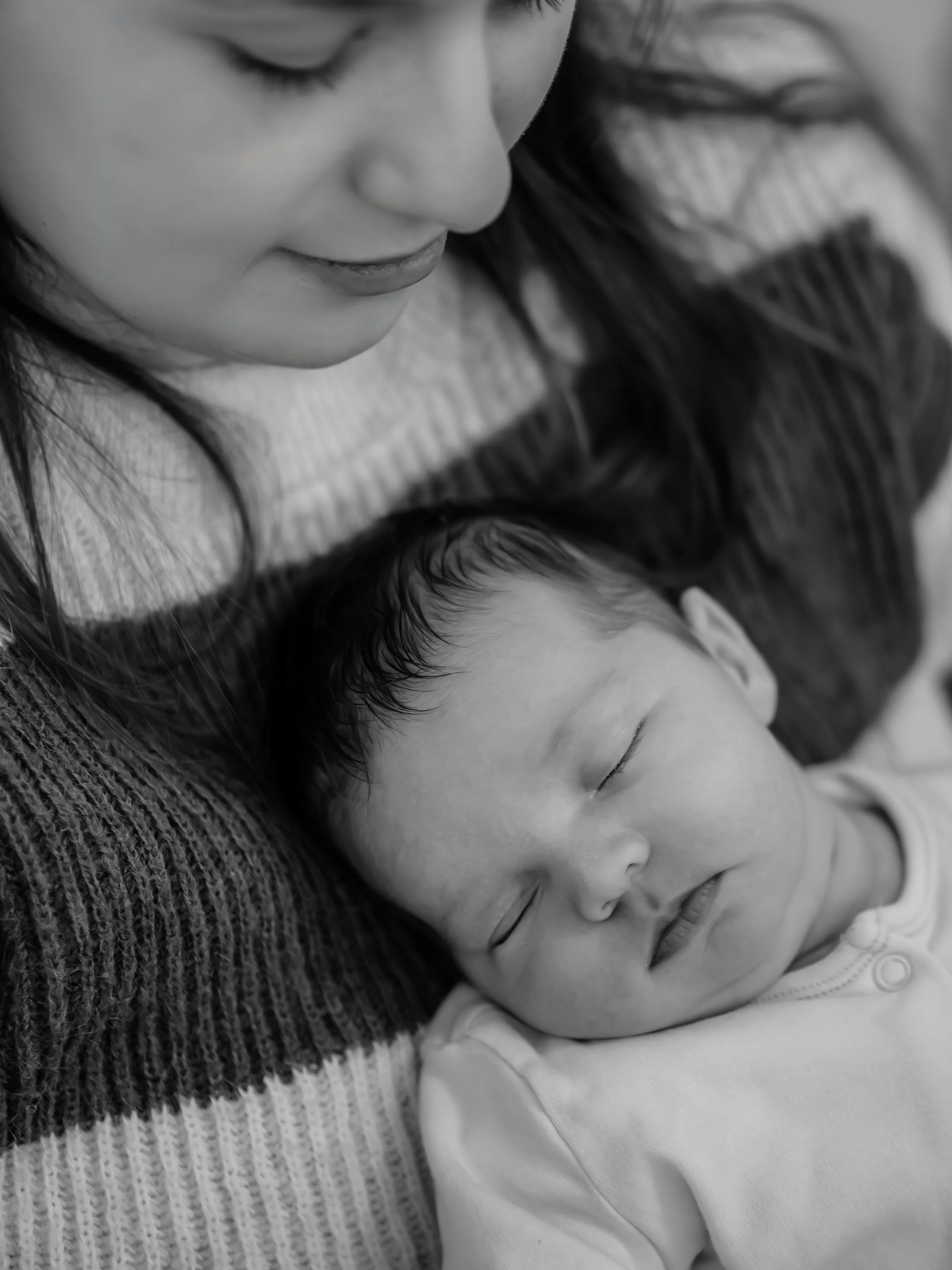 A woman with long dark hair lying down next to a sleeping baby, both resting on a textured blanket, captured in black and white.