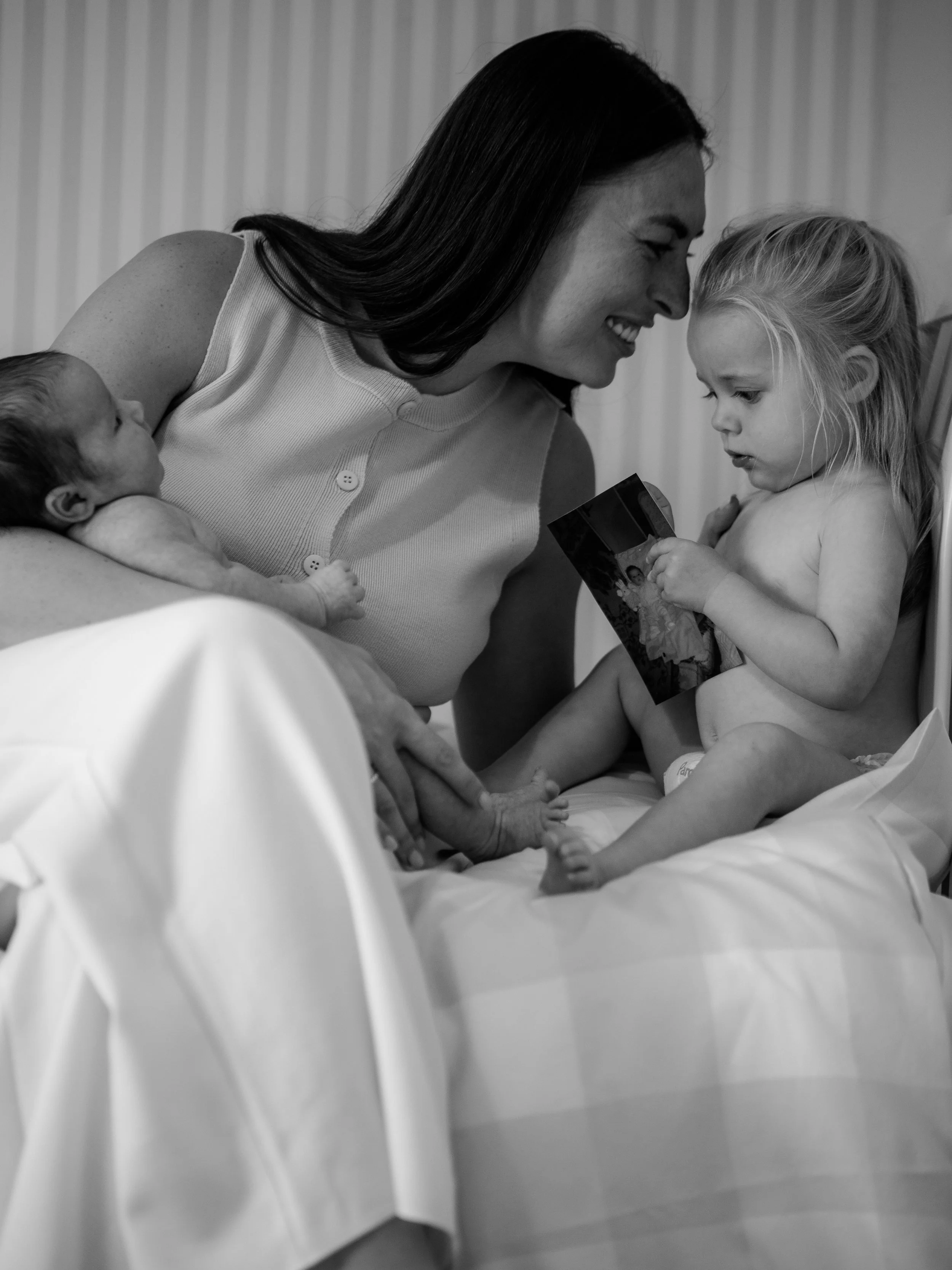 A woman is holding a newborn baby with one arm and a young girl is sitting on a hospital bed looking at a photograph, with the woman smiling and the girl appearing curious.