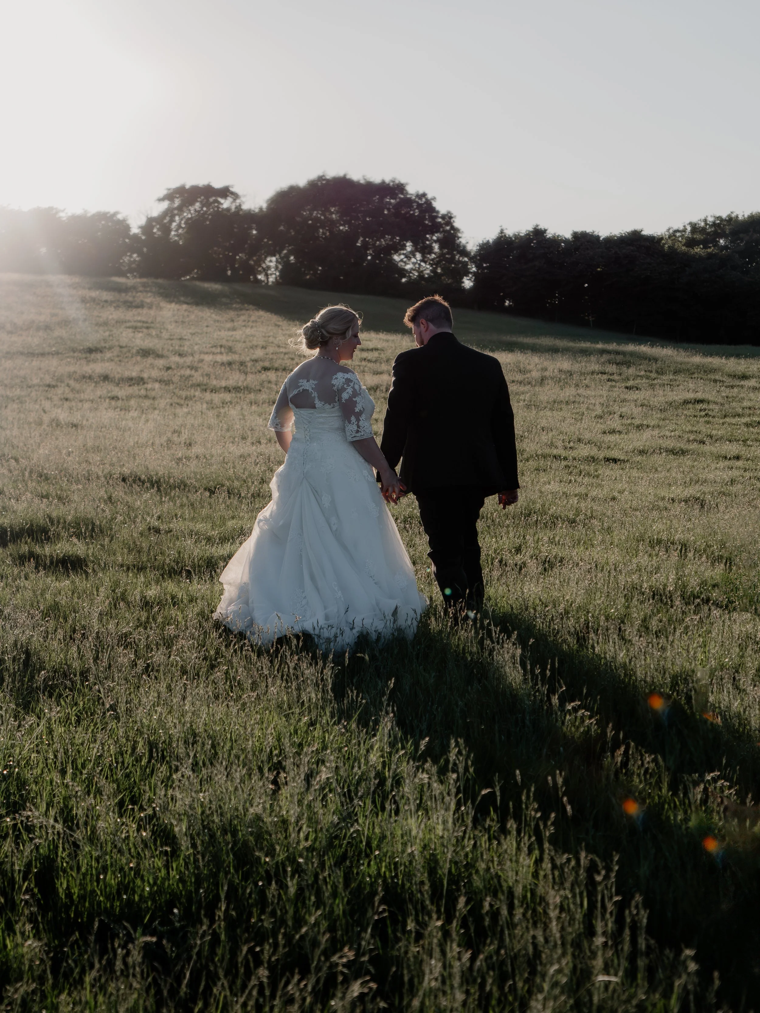 Bride and groom walking hand in hand through a grassy field during sunset.