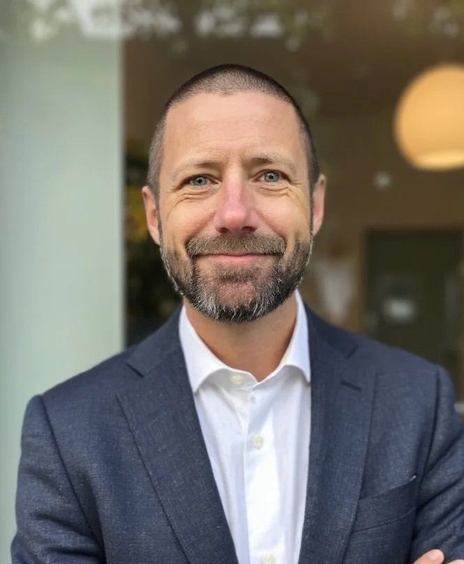 En man med kort hår och skägg, klädd i en mörkblå kostym och vit skjorta, som ler. Bakgrund med fönster och en lampa.