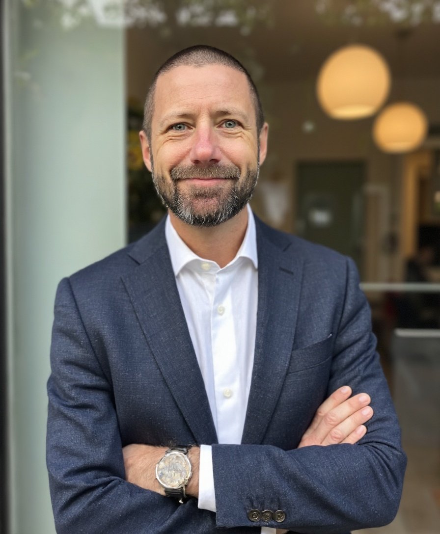 En man i mörk blå kostym och vit skjorta står med armarna korsade framför en bakgrund med moderna lampor och ett fönster.