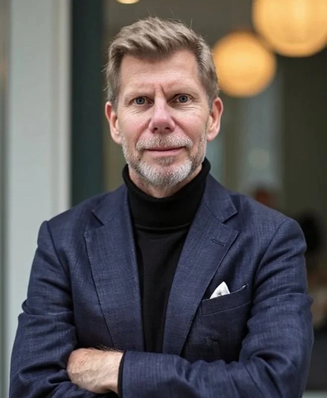 En man med grått hår och skägg, iklädd mörkblå kostym och svart turtleneck, står med korsade armar.