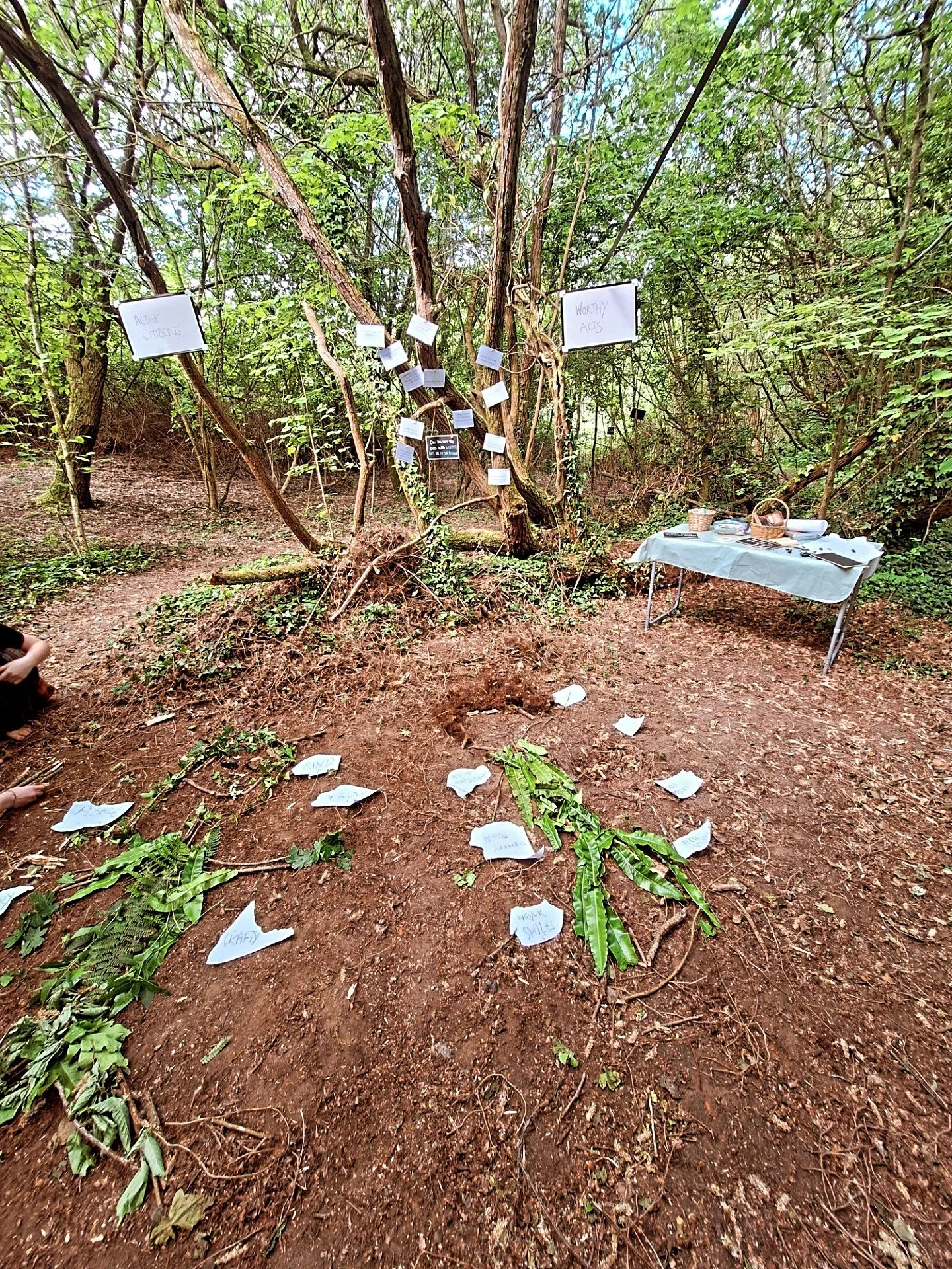 A forest scene with a makeshift display for a community or environmental event, featuring handwritten notes on paper, some attached to trees and others scattered on the ground, along with a small table with supplies on the right side.