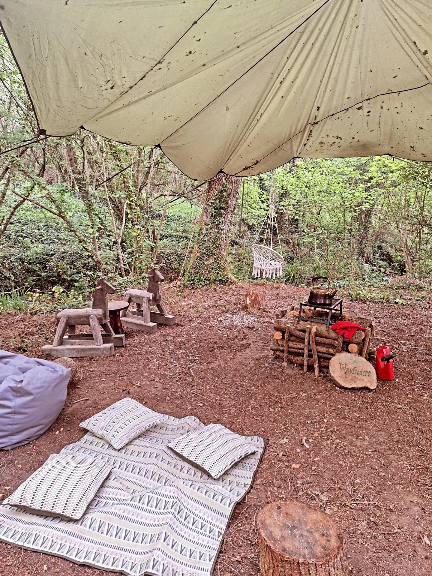 Camping scene in a forest with a beige canopy overhead. In the foreground, there are two pillows and a blanket on the ground. There are two wooden animal-shaped chairs on the left, a small stack of firewood with a kettle and red cloth on top on the right, and a hanging chair in the background among trees and green foliage.
