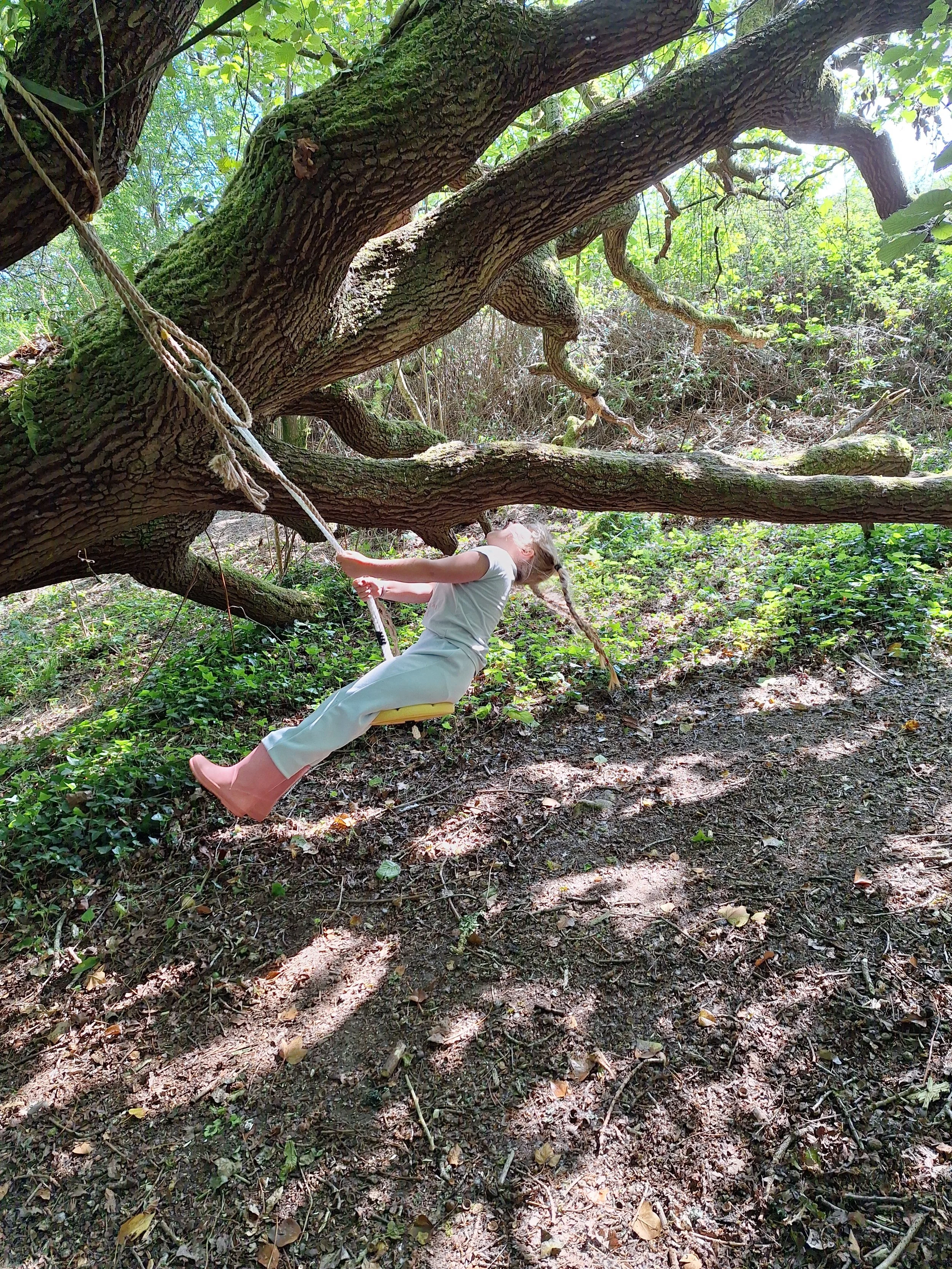 A young girl wearing pink rain boots and light-colored clothes is sitting in a wooden swing tied to a large tree in a forest setting. The girl is leaning back with her head tilted upward, enjoying the outdoors on a sunny day.