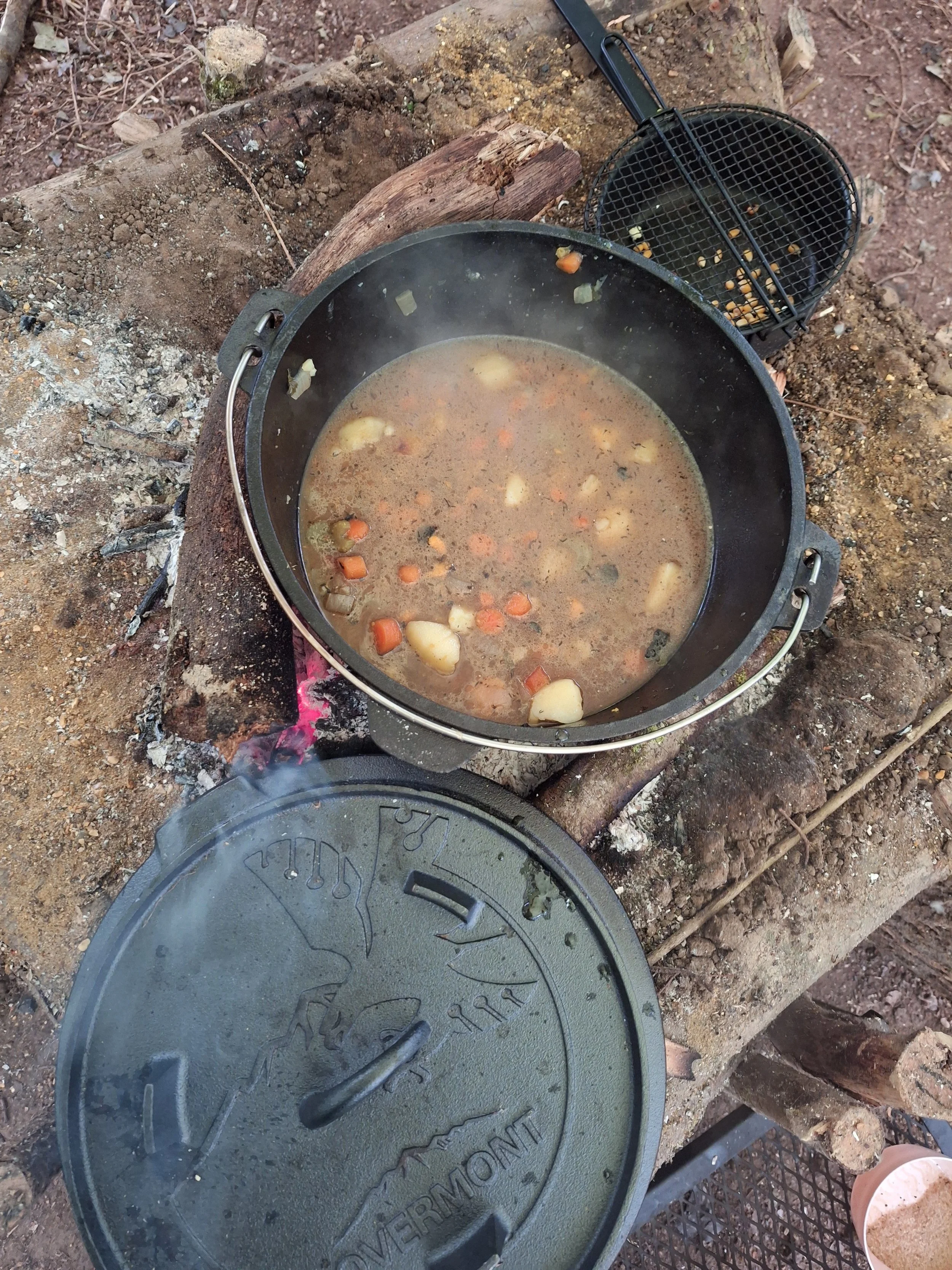 A cast iron Dutch oven on a campfire with a lid partially removed. Inside the Dutch oven, there is a stew with vegetables such as potatoes, carrots, and celery cooking outdoors on a rustic fire pit surrounded by dirt and logs.