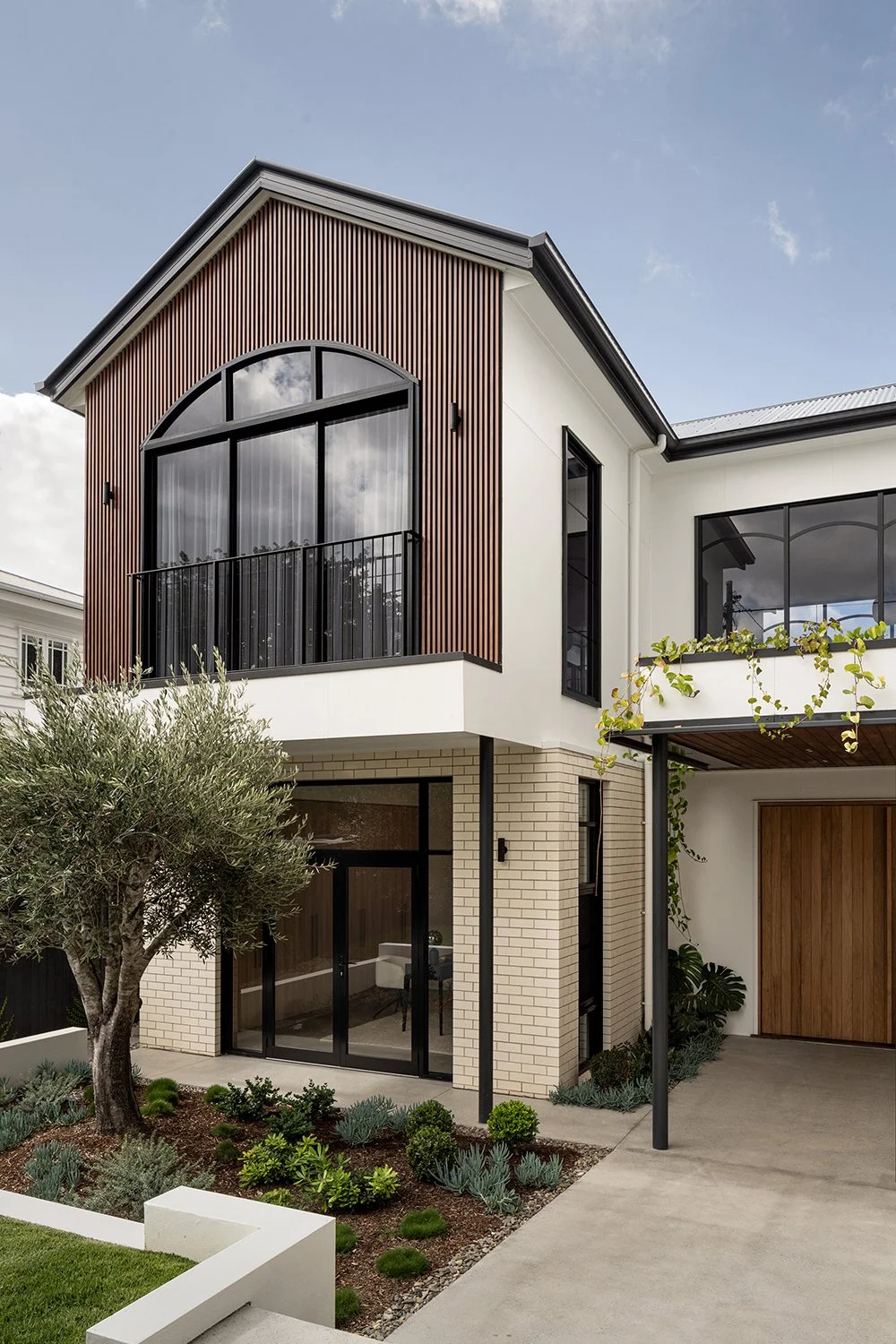 Modern house with a combination of white brick, black accents, and wooden siding, featuring a small front garden with various plants and an olive tree.