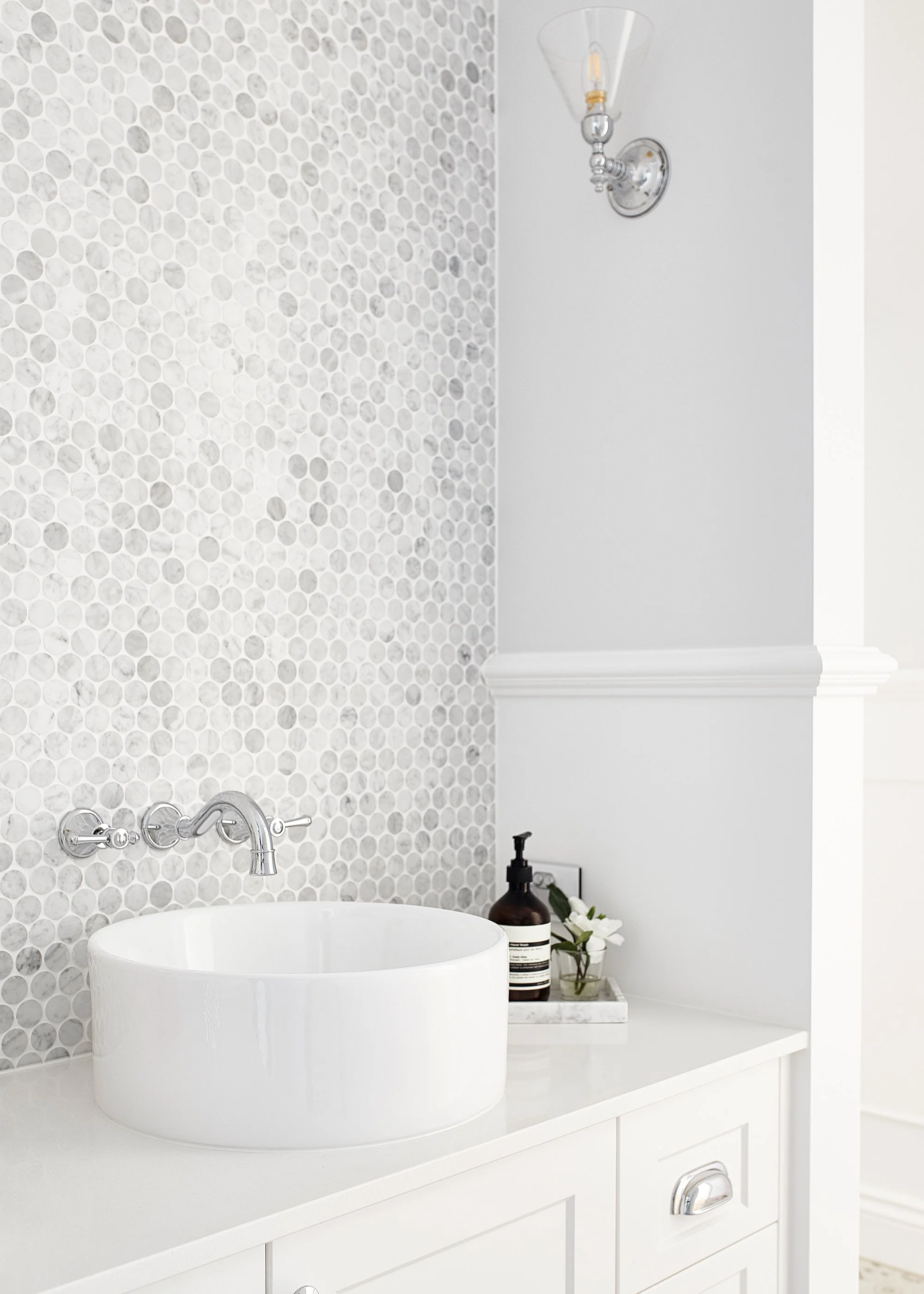 A white bathroom vanity with a round vessel sink, a wall-mounted faucet, and a black soap dispenser with flowers on a tray. There's a textured tile backsplash and a wall sconce light above.