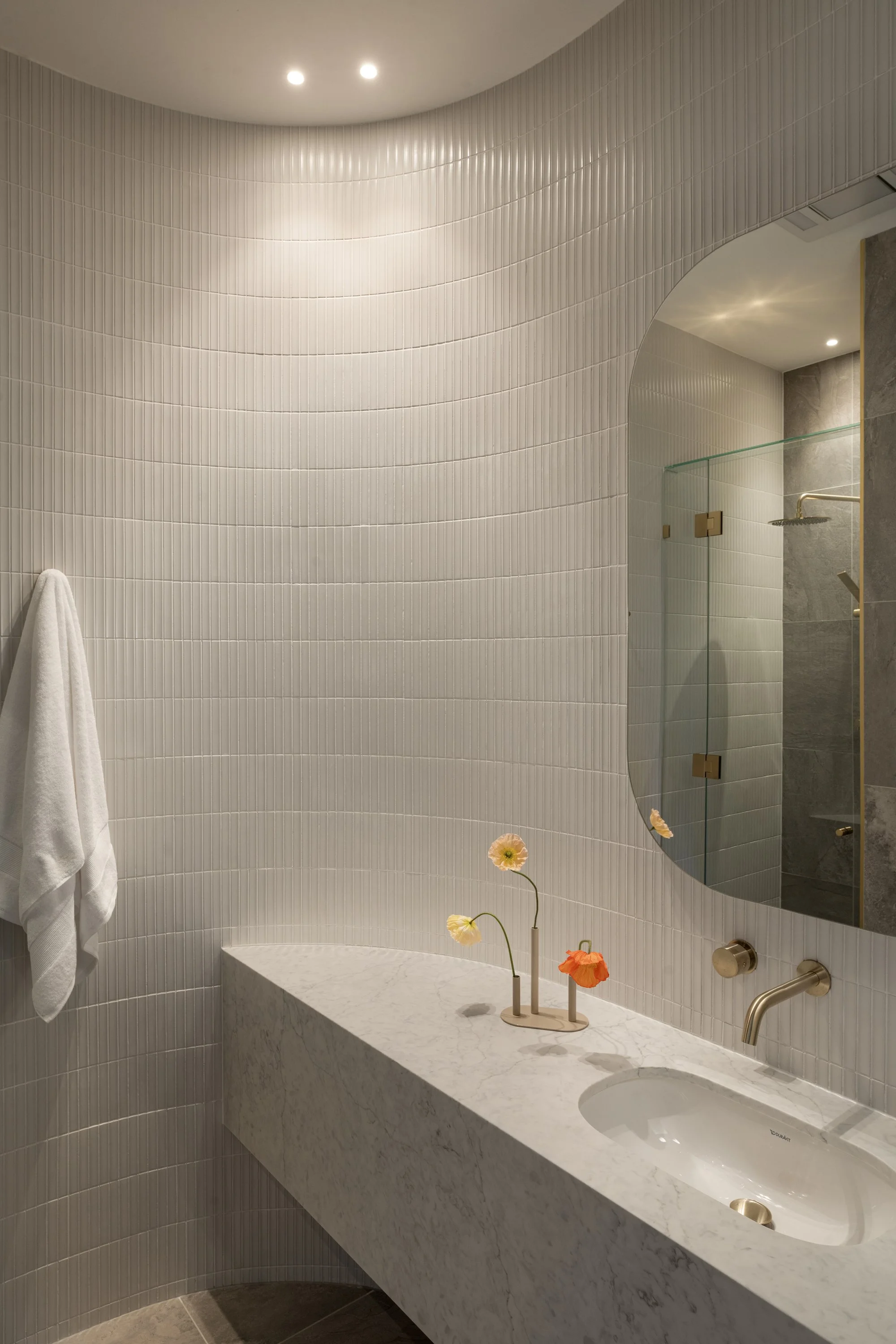 Modern bathroom with curved tiled wall, marble countertop sink, and a large mirror. Small vase with colorful flowers on the counter, with a towel hanging on the wall.