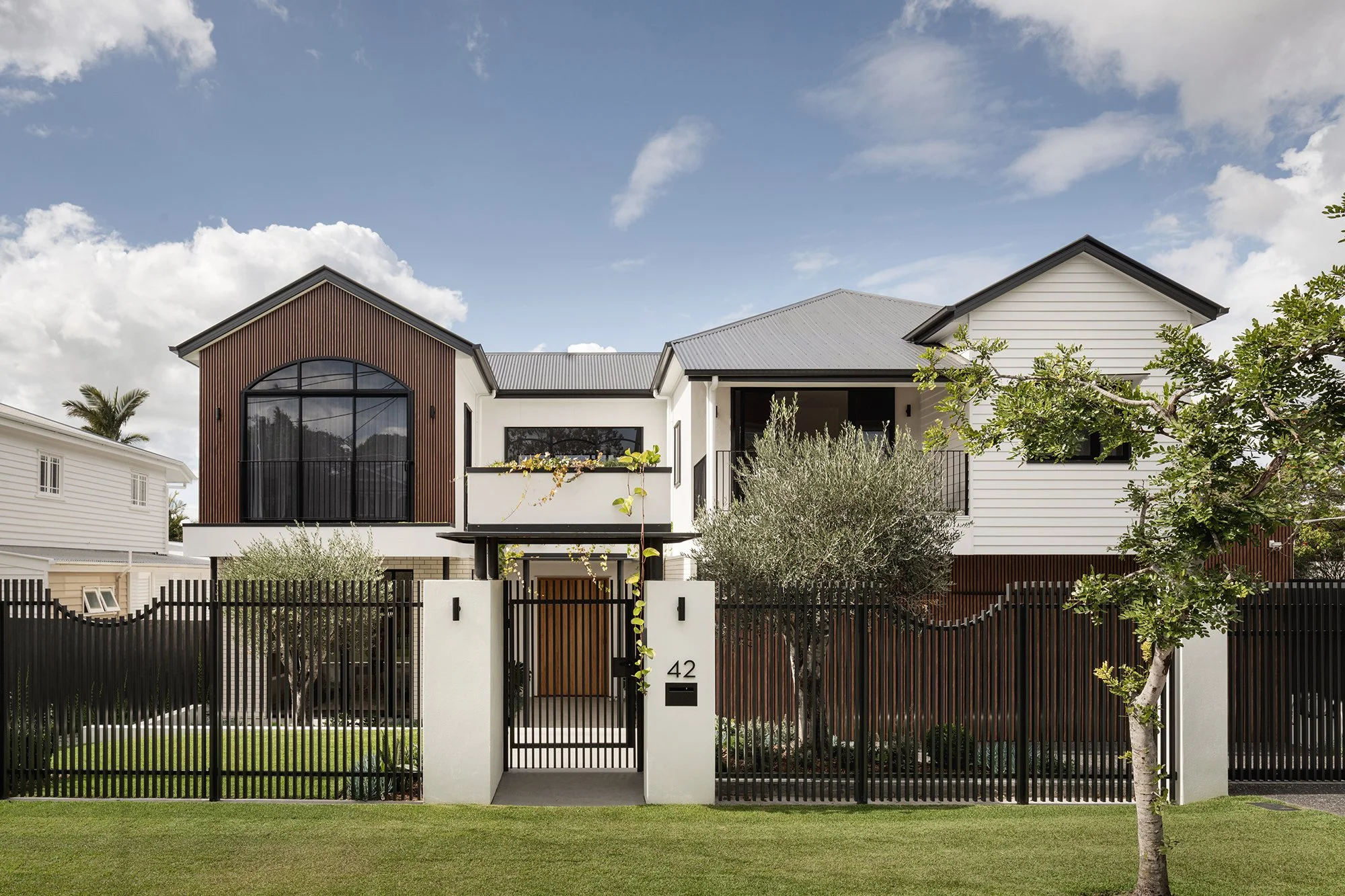 Modern two-story house with black fence, gated entrance, and well-maintained lawn; white and wood exterior, large windows, and separated sections under a partly cloudy sky.