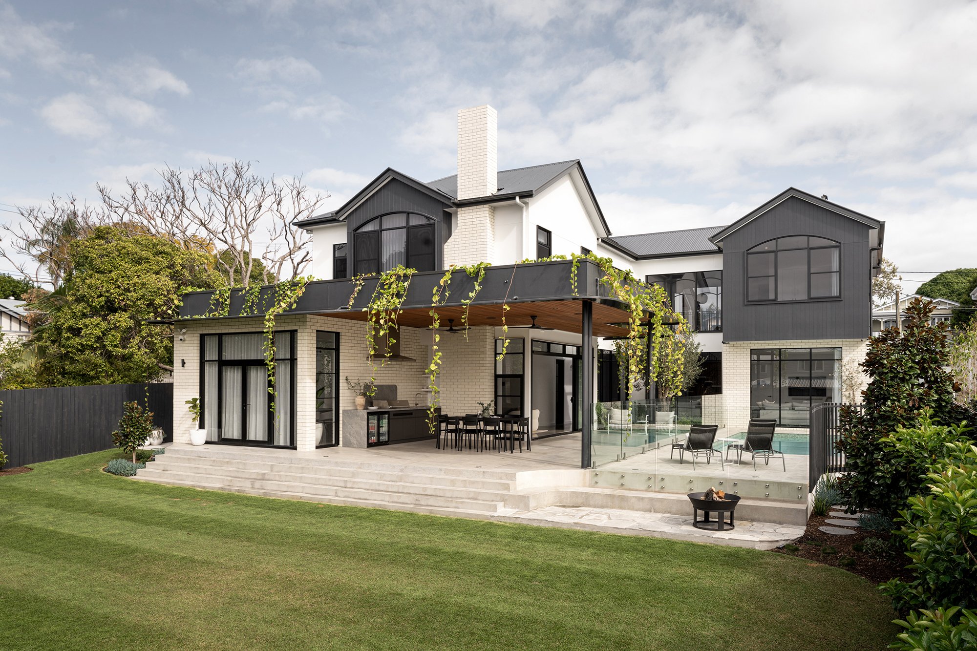 Modern two-story house with a backyard patio, outdoor dining area, lounge chairs, a fire pit, and a small swimming pool surrounded by a glass fence, greenery, and trees.