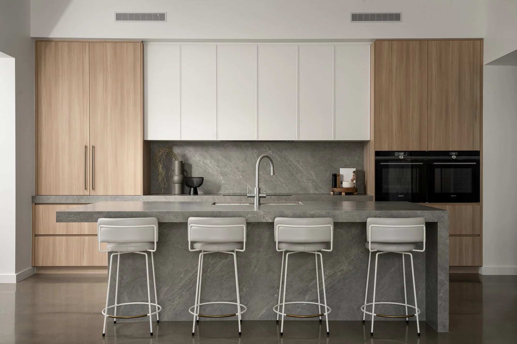 Modern kitchen with light wood and white cabinetry, gray stone countertops and backsplash, four white barstools at a gray stone island, built-in black appliances.